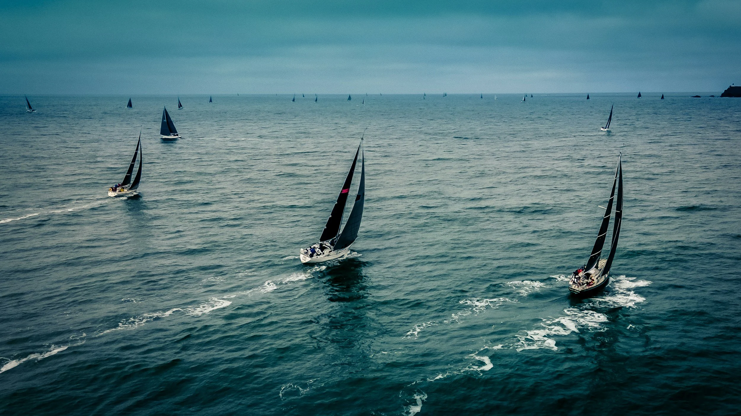 Multiple sailboats racing on open water under cloudy sky.