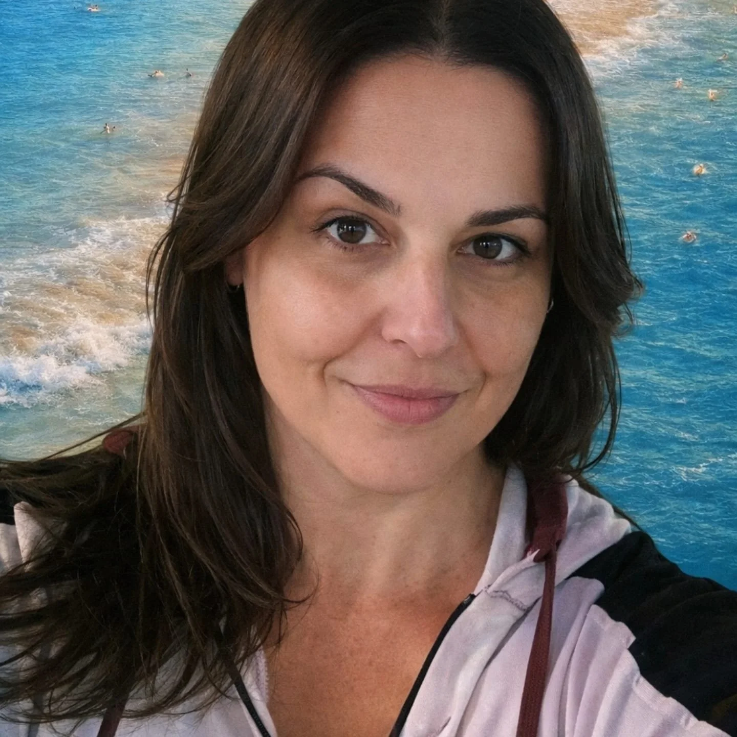 Mulher com cabelo castanho escuro, rosto sorridente e fundo de mar com ondas e pessoas na água.