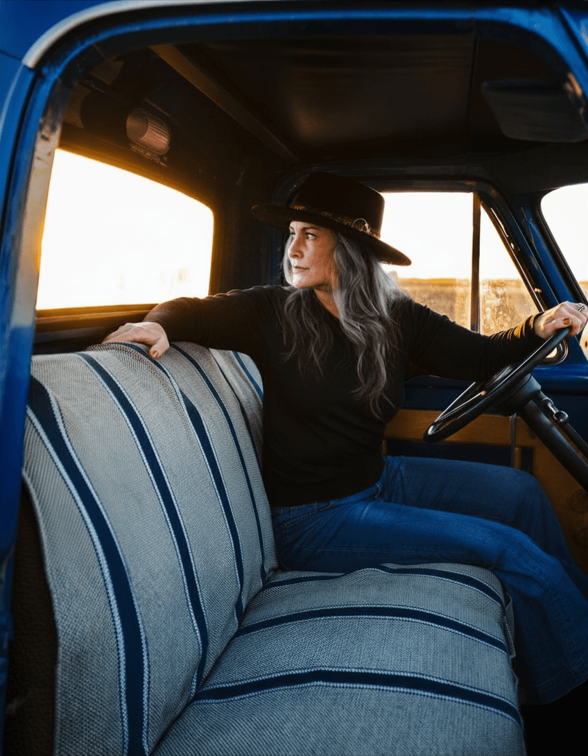 A woman with gray hair wearing a black hat and black shirt sitting in the driver's seat of a vintage blue truck with striped upholstery, looking out the window during sunset.