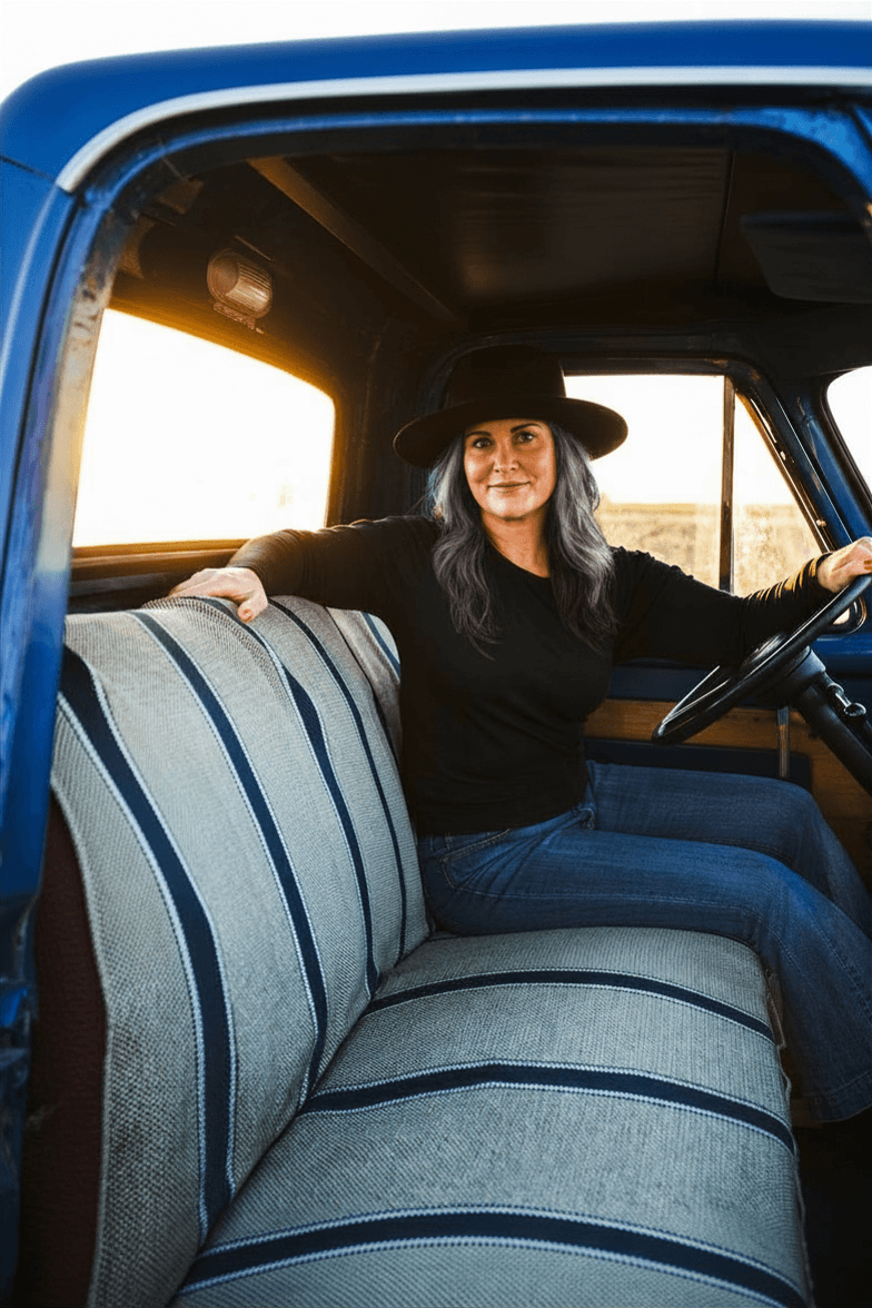 Woman sitting in the driver's seat of a vintage blue truck, wearing a black hat and black long-sleeve shirt, with a sunset in the background.