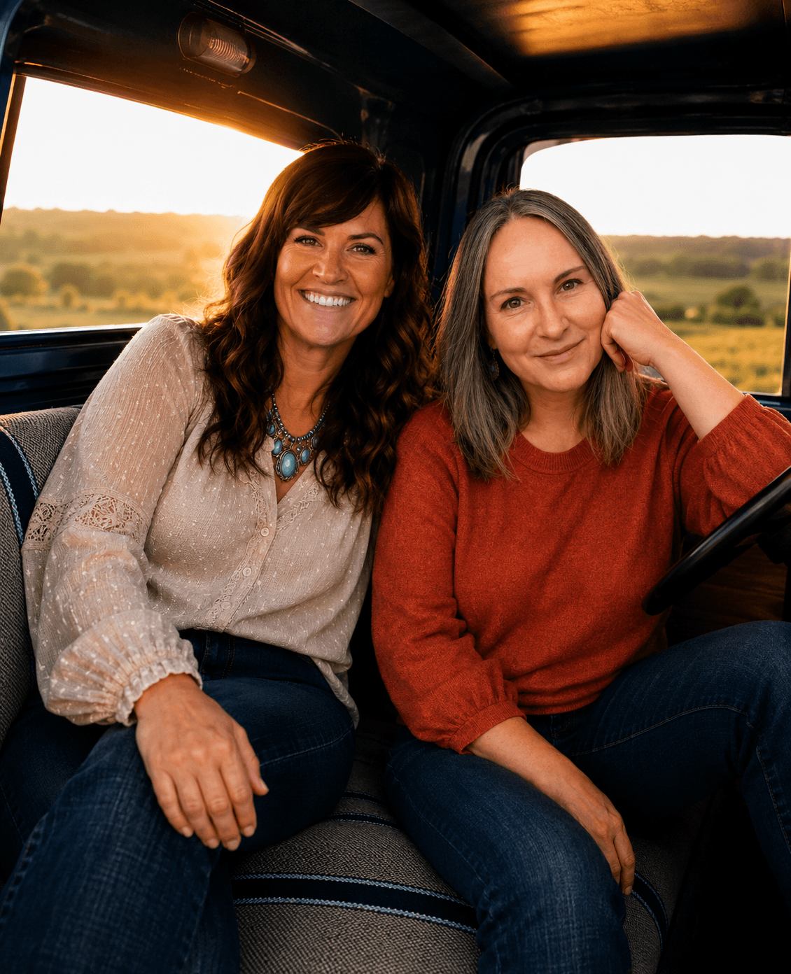 Two women sitting inside a vehicle, smiling at the camera, with a sunset view of a rural landscape outside the window.