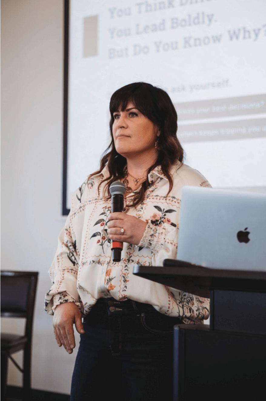 A woman with dark hair standing in front of a presentation screen, holding a microphone. She wears a floral-patterned blouse, and there is a silver Apple MacBook on a table beside her.