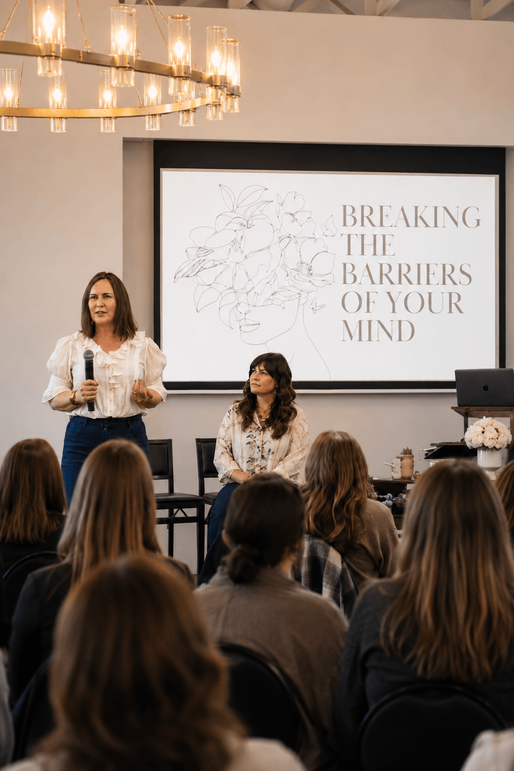 Woman holding a microphone speaking at a seminar or conference, with a seated woman and audience members watching, and a presentation slide behind with the text 'Breaking the Barriers of Your Mind' and a line drawing of a face with flowers.
