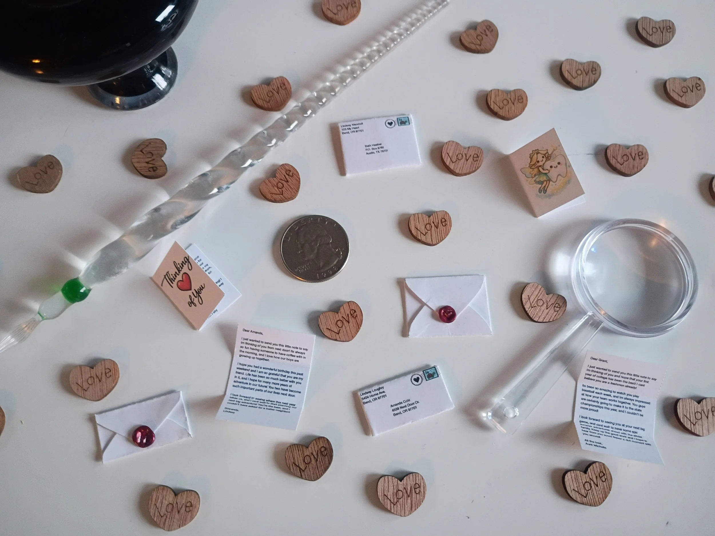 Various small wooden heart-shaped tokens with the word 'Love' engraved on them, tiny envelopes with red wax seals, tiny handwritten notes, a clear glass magnifying glass, an ink well, and a glass calligraphy pen all on a white table.