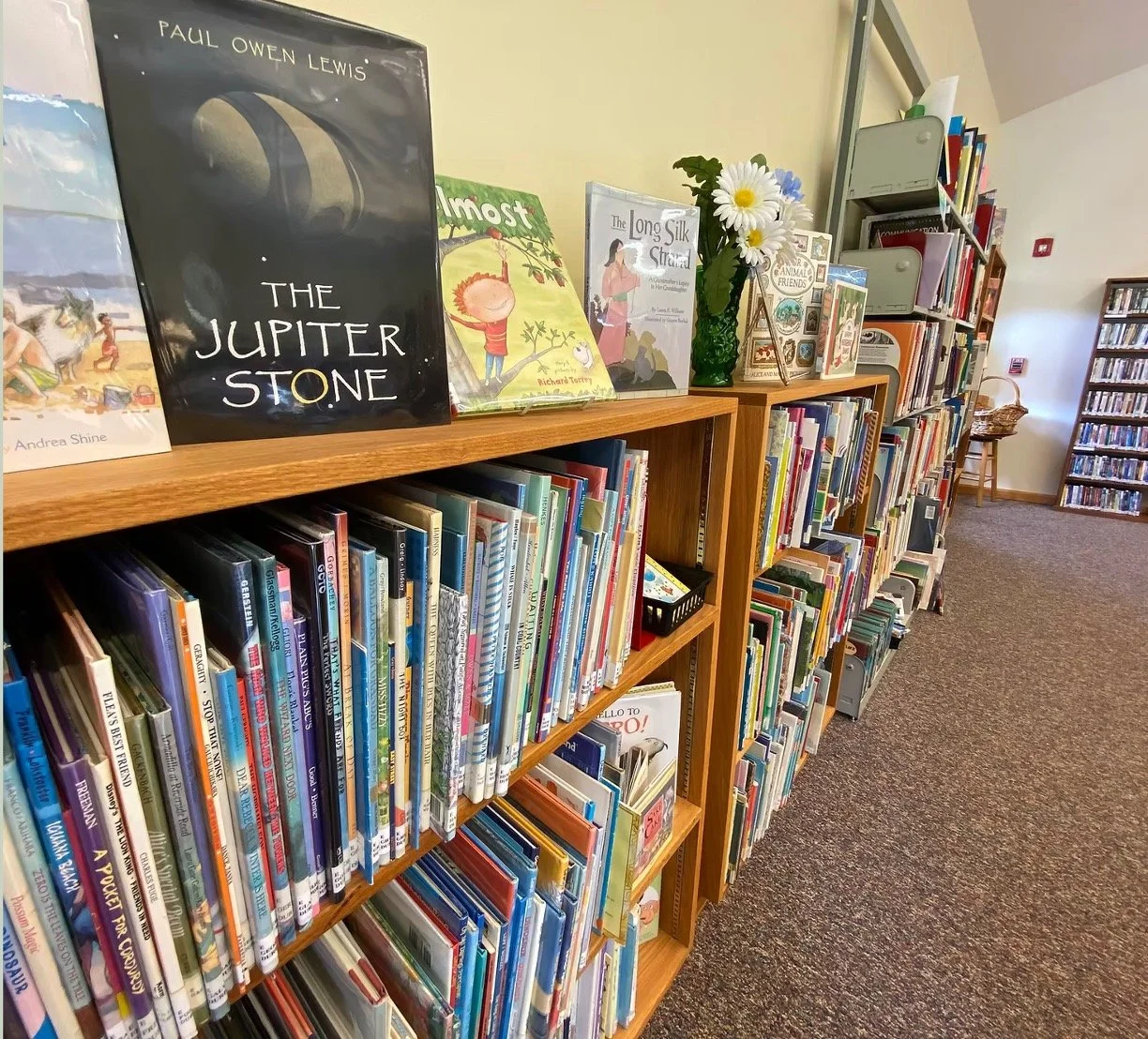 Bookshelves filled with children and young adult books in a library, with some books displayed facing outward, including titles such as "The Jupiter Stone" and "The Long Silk String". A green vase with daisies and other flowers is on top of the shelves, and a small black basket with children's books is also visible.