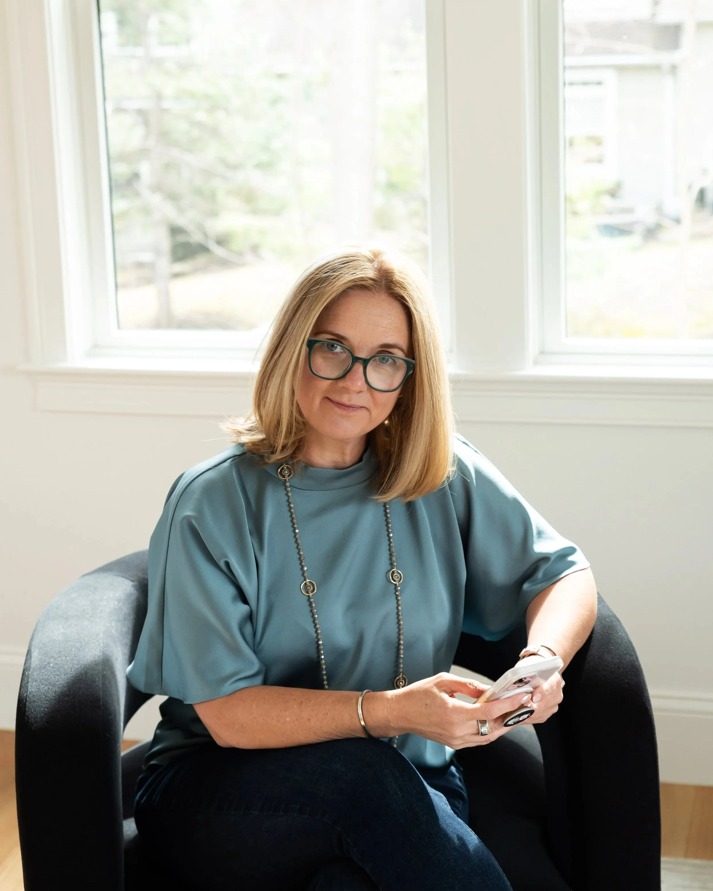 Amy Slate, a woman with blonde hair, glasses, and a blue top, sitting in a black chair and looking at the camera indoors, with windows and a view of trees outside.