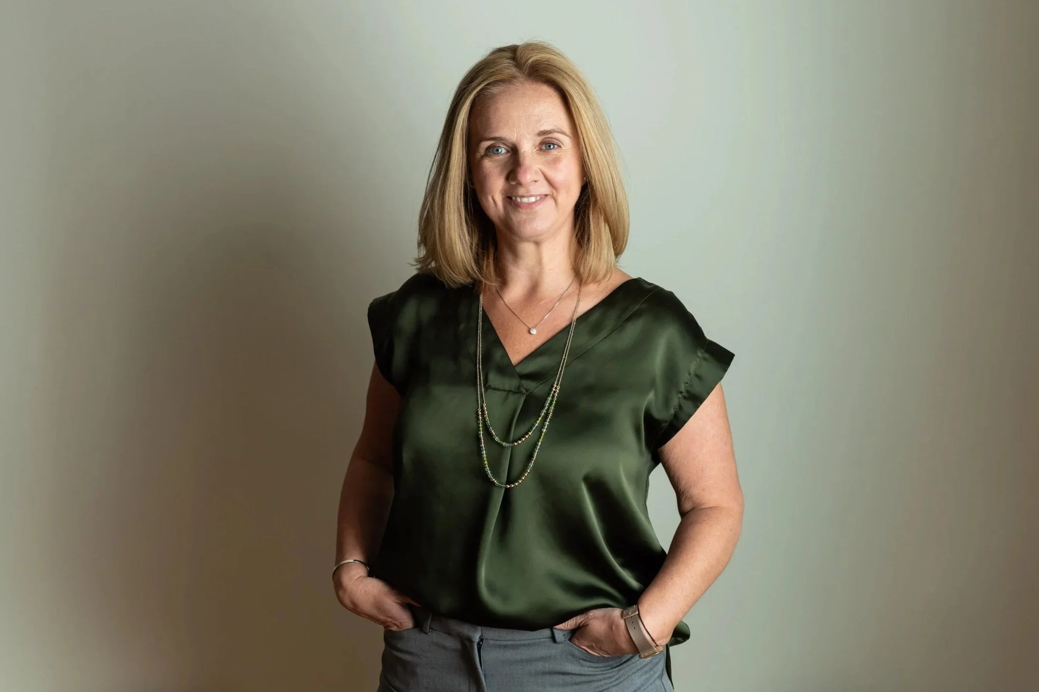 Amy Slate, the Breakup Broker, a woman with blonde hair, wearing a dark green satin blouse and gray pants, standing with her hands in her pockets against a plain light-colored wall.