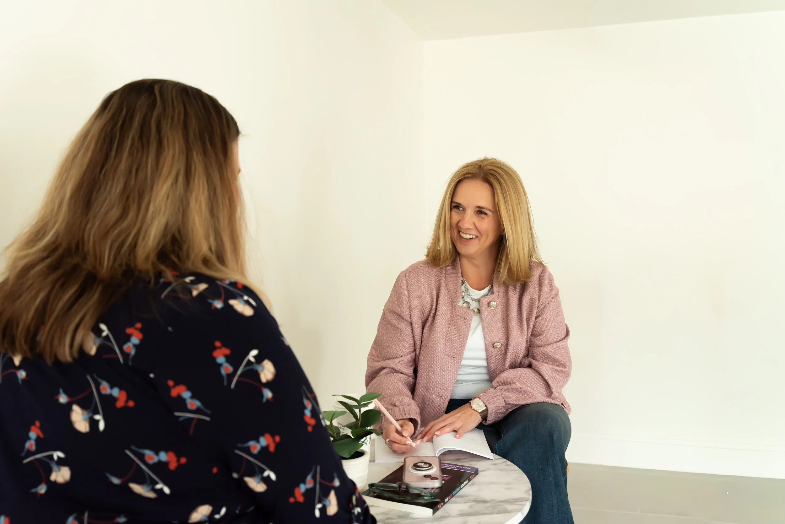 Two women having a conversation in a bright, minimalistic room. One woman is facing away from the camera, wearing a dark floral top, and the other woman is facing the camera, wearing a pink jacket and smiling.