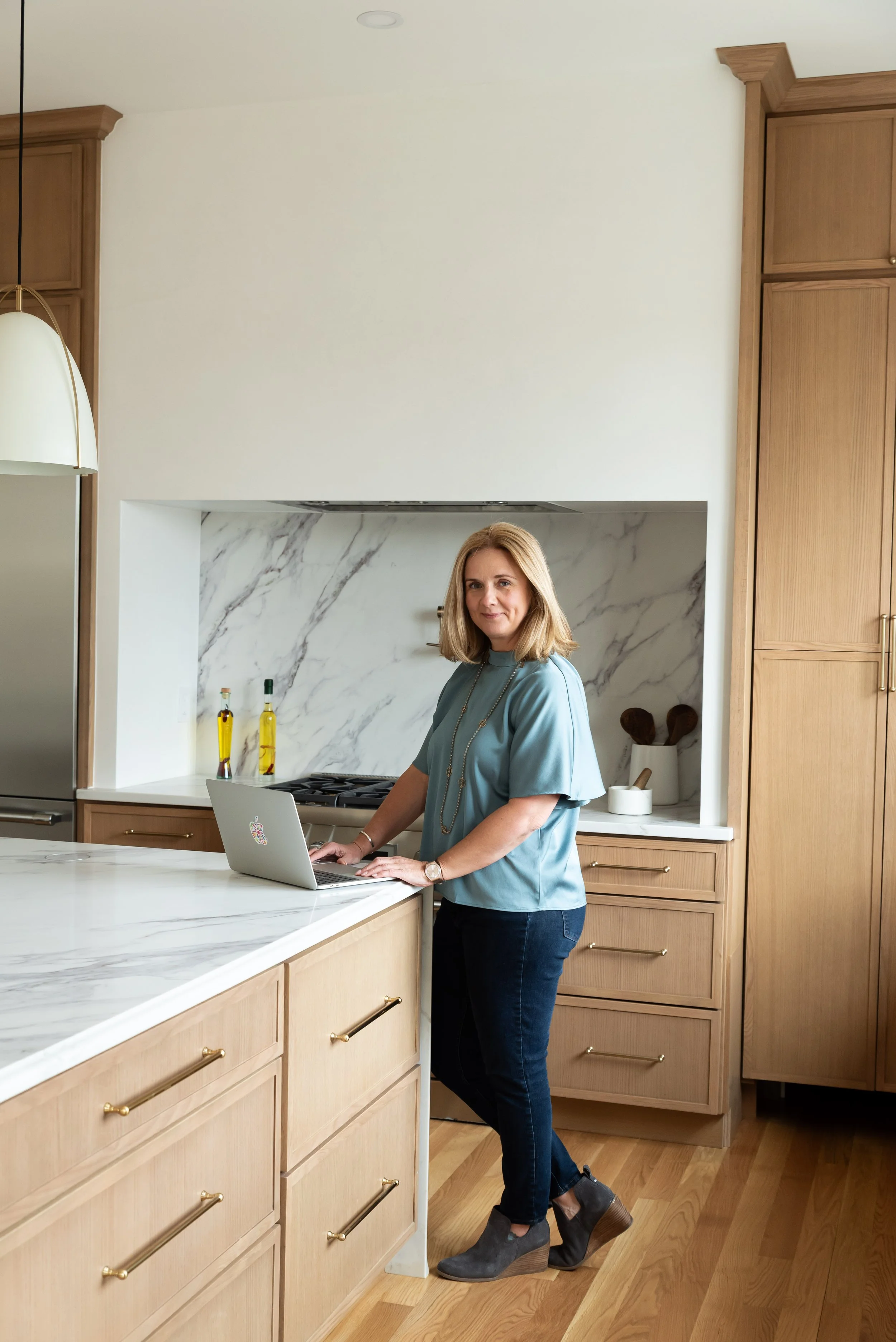 Amy, the Breakup Broker, with blonde hair in a short bob, wearing a teal blouse, dark jeans, and gray ankle boots, standing in a modern kitchen with a marble countertop, wooden cabinets, and a laptop open in front of her.