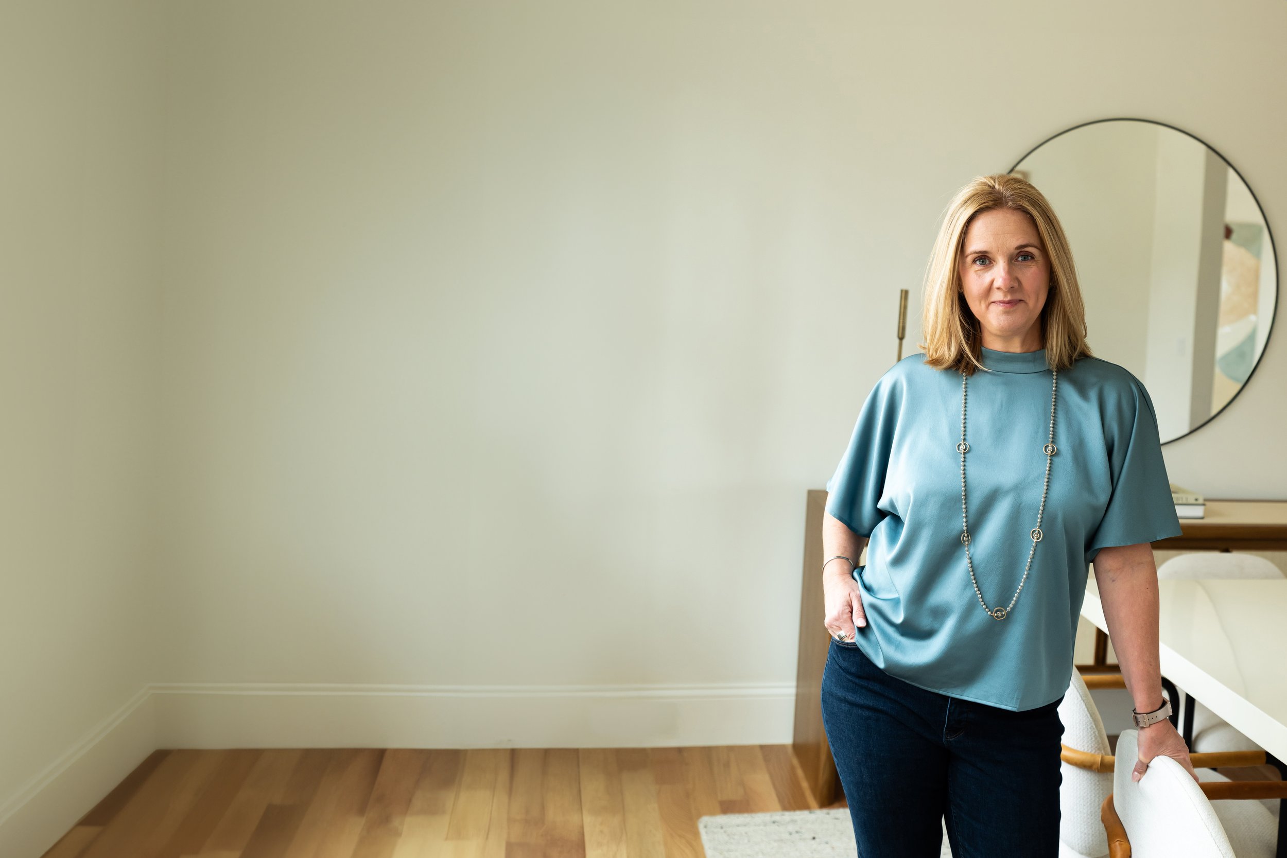 A woman with shoulder-length blonde hair standing in a modern living room, wearing a blue satin blouse with a long necklace, dark jeans, and a watch, looking at the camera.