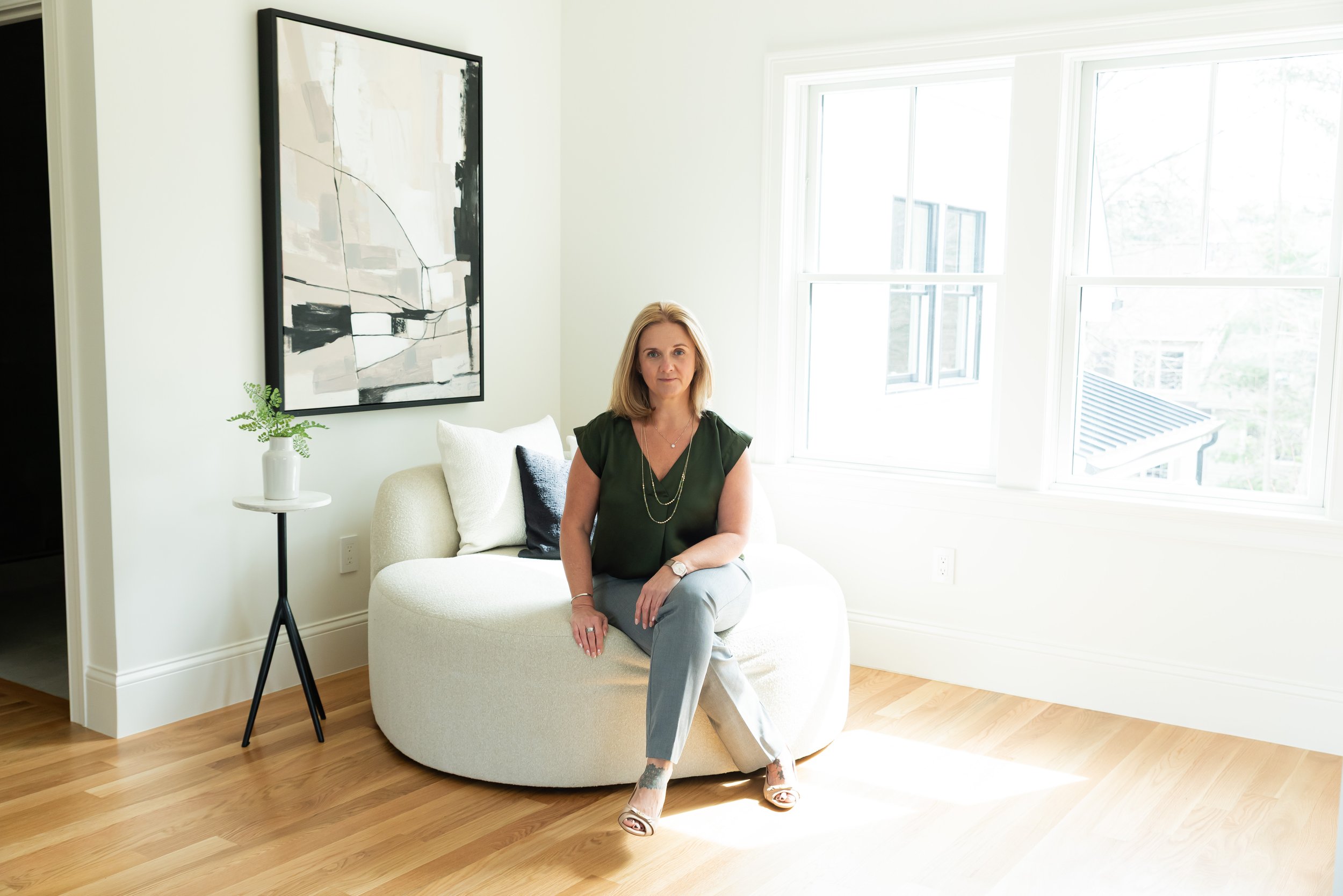 A woman in a green top and light-colored pants sitting on a white, rounded sofa in a bright living room with large windows and wooden floors. There is a black and white abstract painting on the wall and a small white table with a plant.