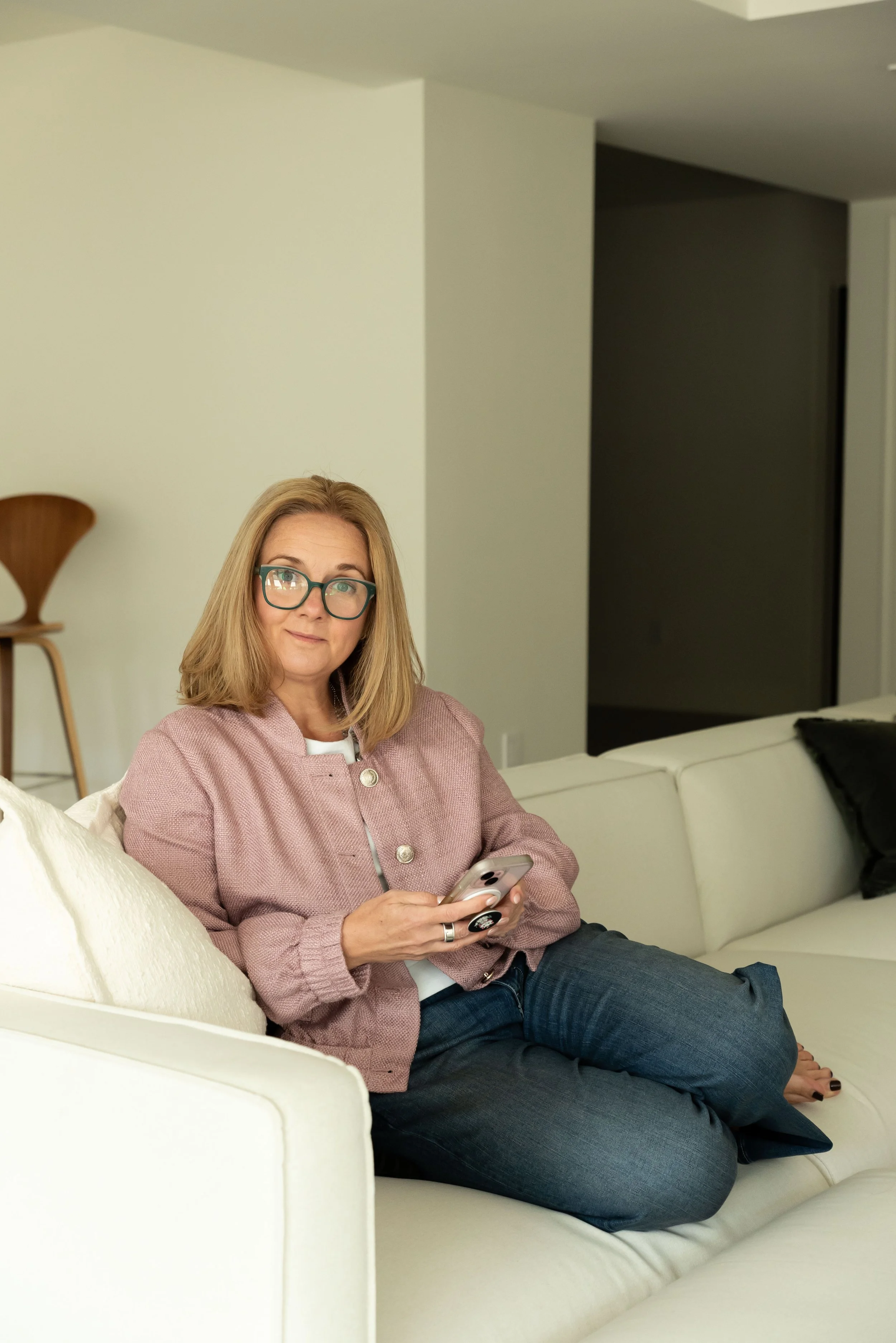 A woman with blonde hair, glasses, wearing a pink jacket, sitting on a white sofa, holding a smartphone.
