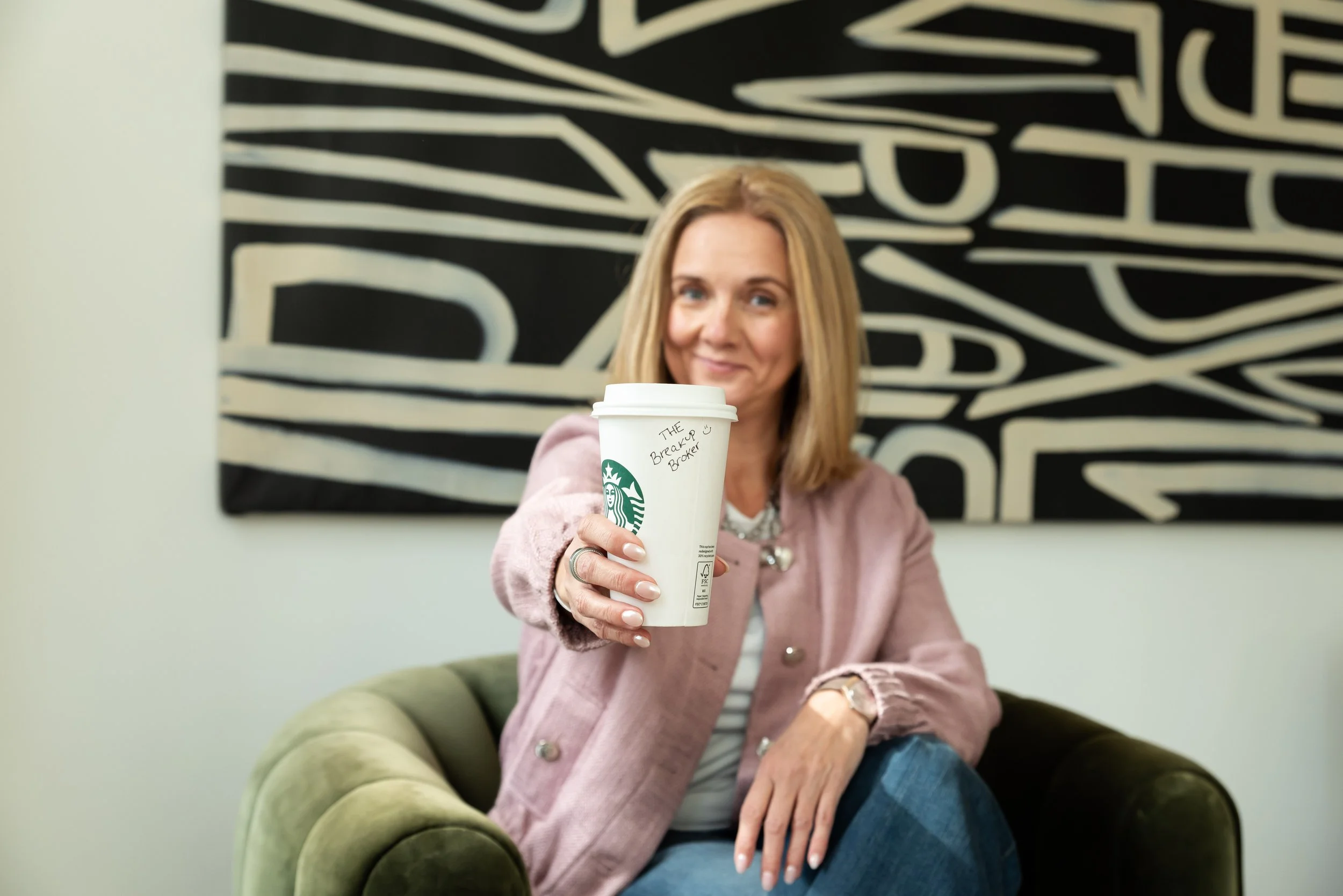 Woman sitting on a green armchair, holding a Starbucks coffee cup toward the camera with a note that reads 'The Breaker Ubreaker', smiling, with abstract black and white art in the background.