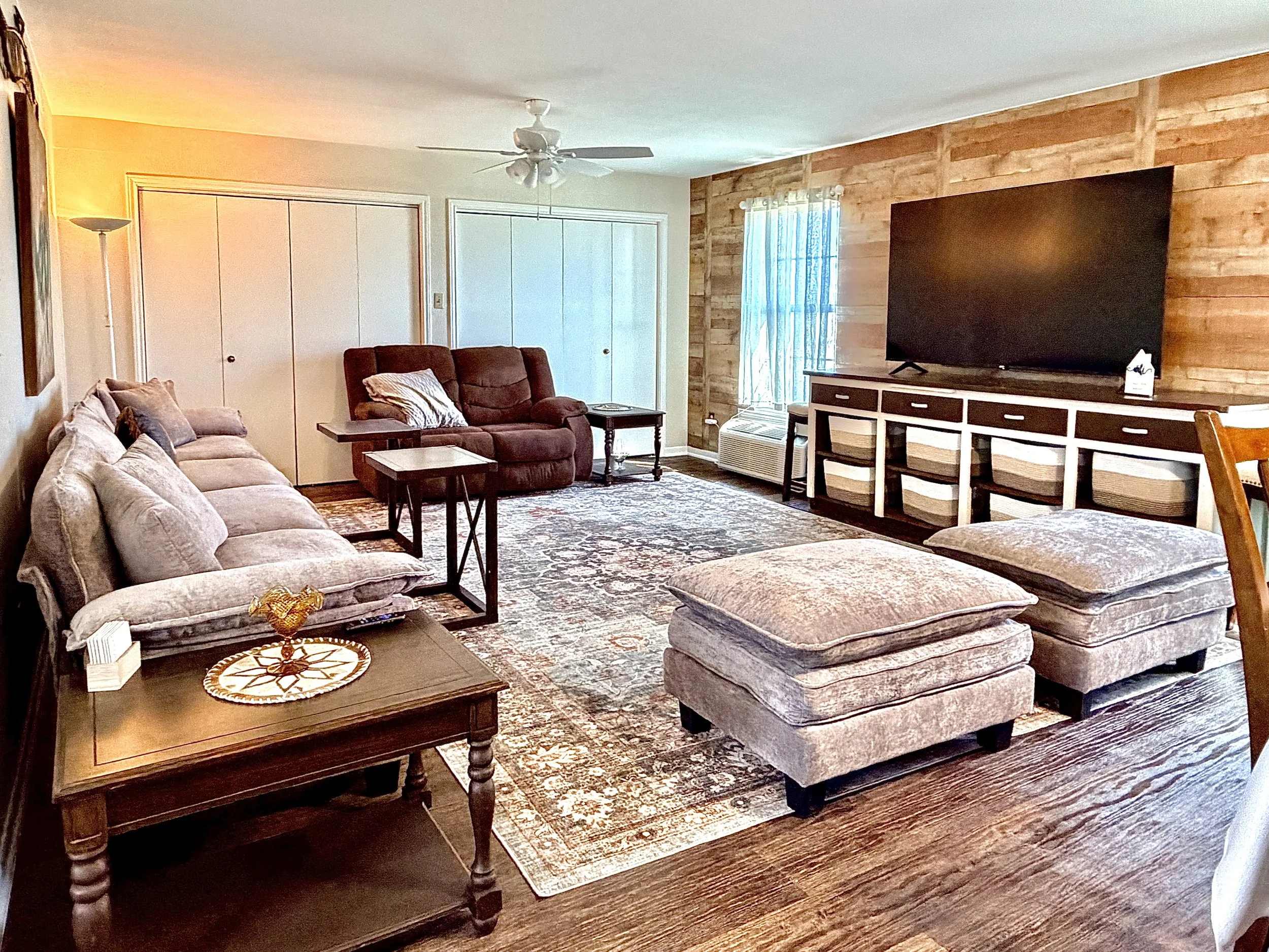 A living room with a beige sofa, two matching ottomans, a burgundy recliner, wooden side tables, and a large flat-screen TV on a patterned wall. The room has a wood-paneled accent wall, a window with white curtains, and a decorative area rug.