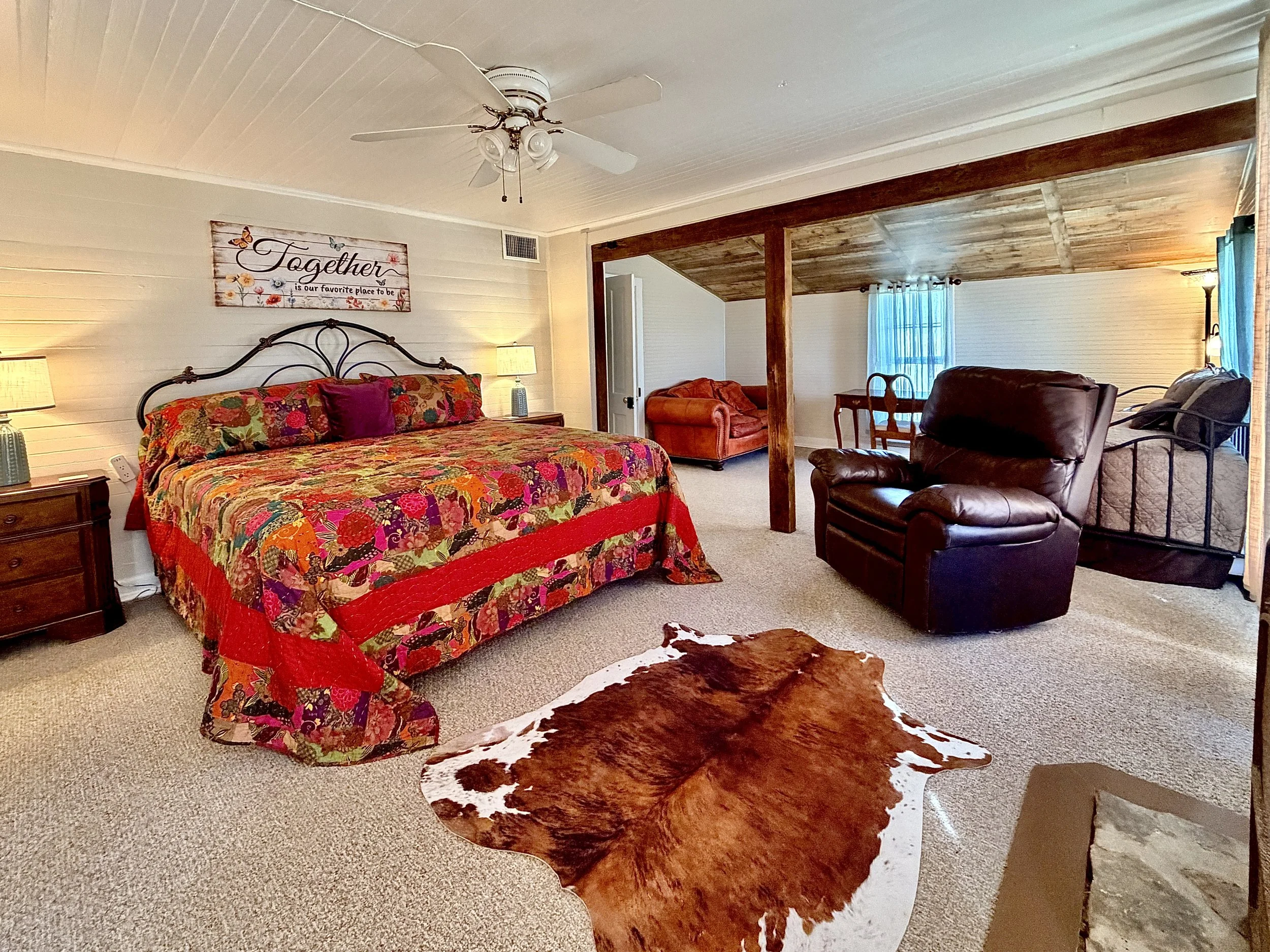 A bedroom with a metal bed frame, patterned quilt, and matching pillows. Wood-paneled wall with a horse portrait above the bed. Two bedside tables with oil lamps, and small windows on side walls, with carpeted floor.