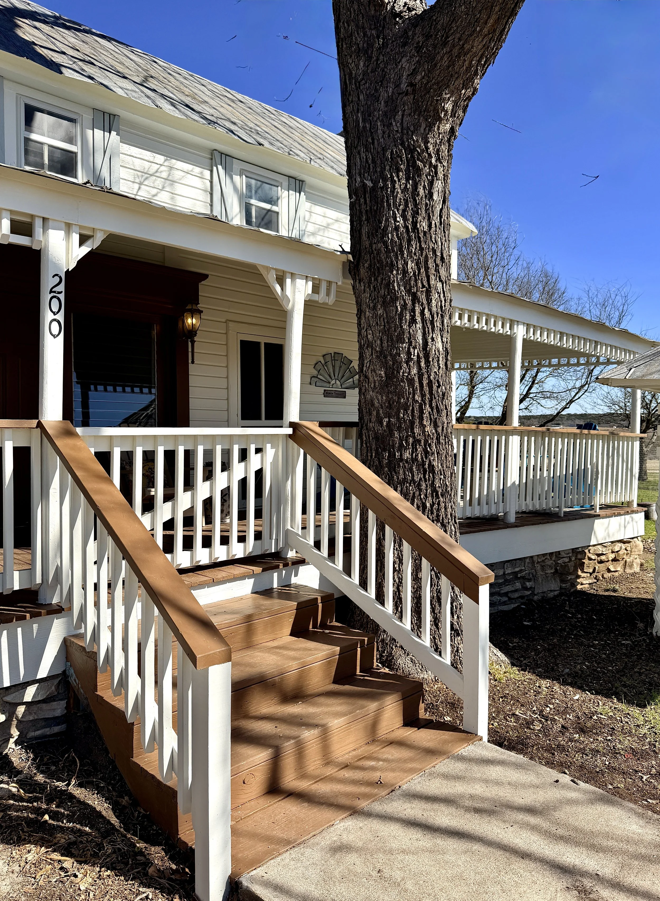 Front porch of a white, two-story house with a wooden staircase, railing, and a large tree in front. The house features a covered porch and a gabled roof, with windows and a door visible.
