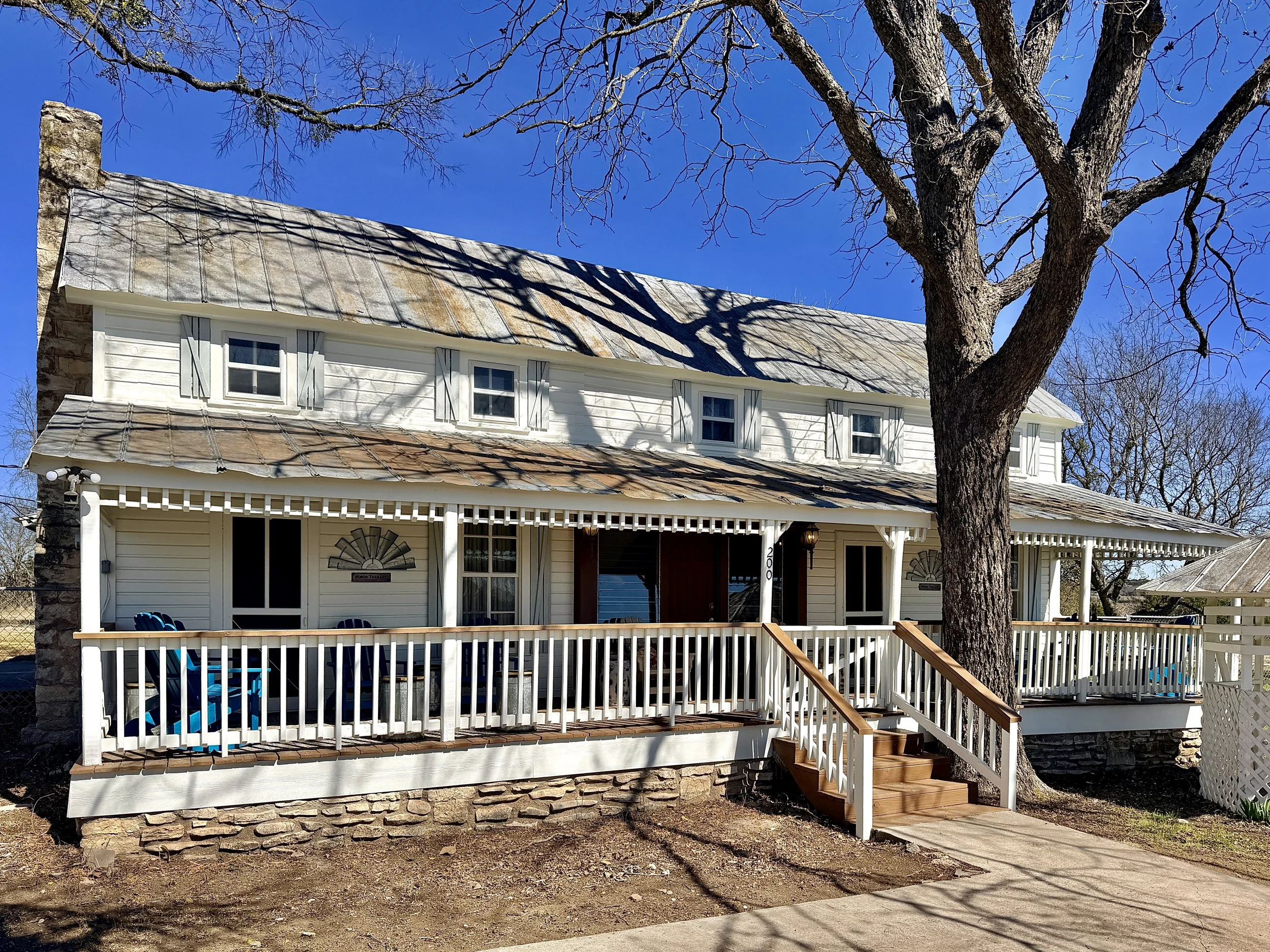 A two-story house with white siding and a stone foundation, featuring a front porch with white railing, steps, and blue chairs. A large tree in the front yard casts shadows on the house. The sky is clear and blue.