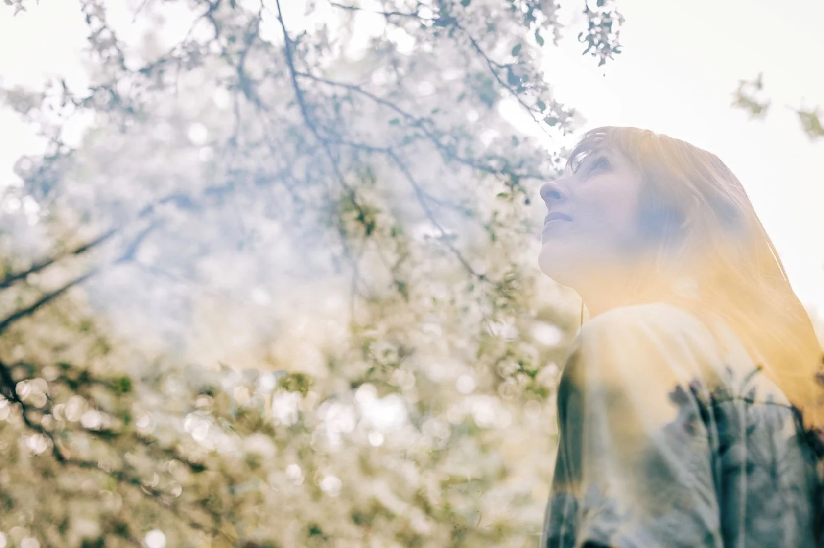 A young woman with long red hair looking upwards, outdoors during daytime with sunlight filtering through tree branches and leaves.