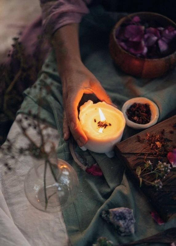 A person holding a lit candle over a picnic setup with bowls of herbs and flowers.