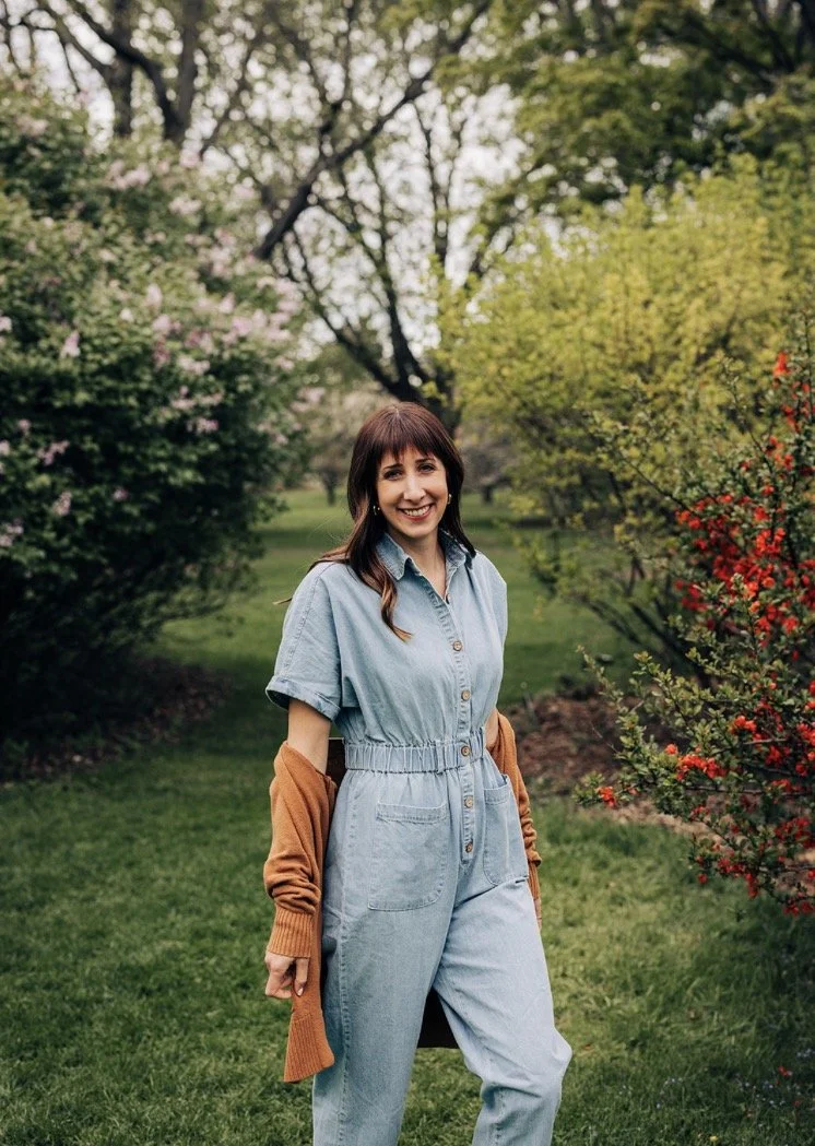 A woman standing in a park or garden with green grass, trees, and blooming bushes, smiling at the camera.