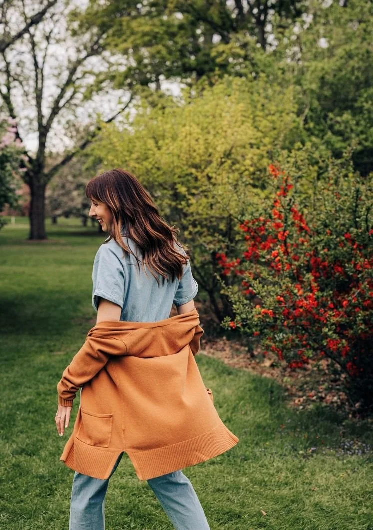 A woman with wavy brown hair smiling and walking in a park with green grass and trees with green leaves, wearing a light blue t-shirt, light blue jeans, and a tan oversized cardigan.