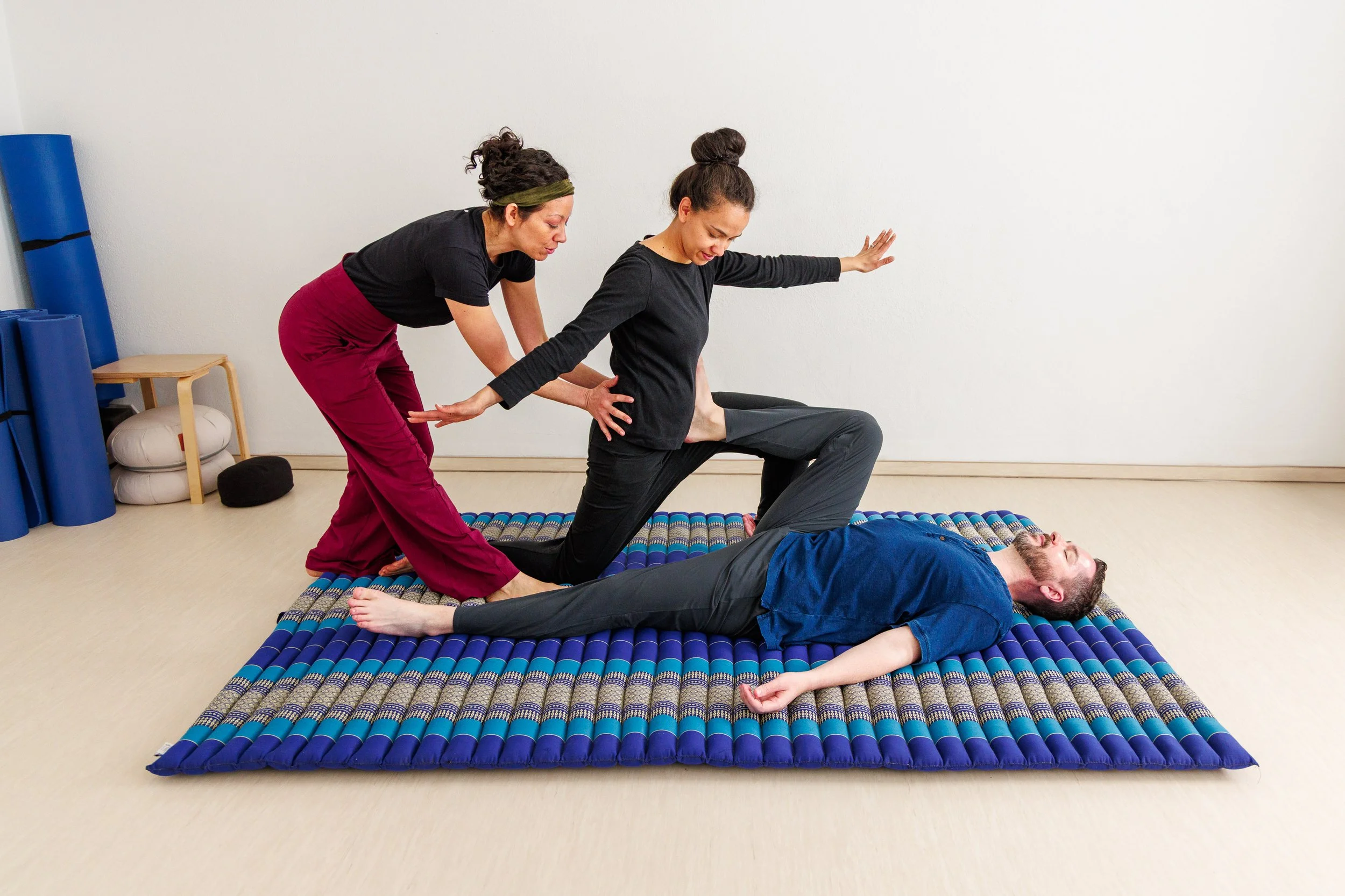 Zwei Frauen helfen einem Mann bei einer Yoga-Übung auf Matte. Der Mann liegt auf dem Rücken, eine Frau unterstützt seinen Oberkörper, die andere seine Hüfte.