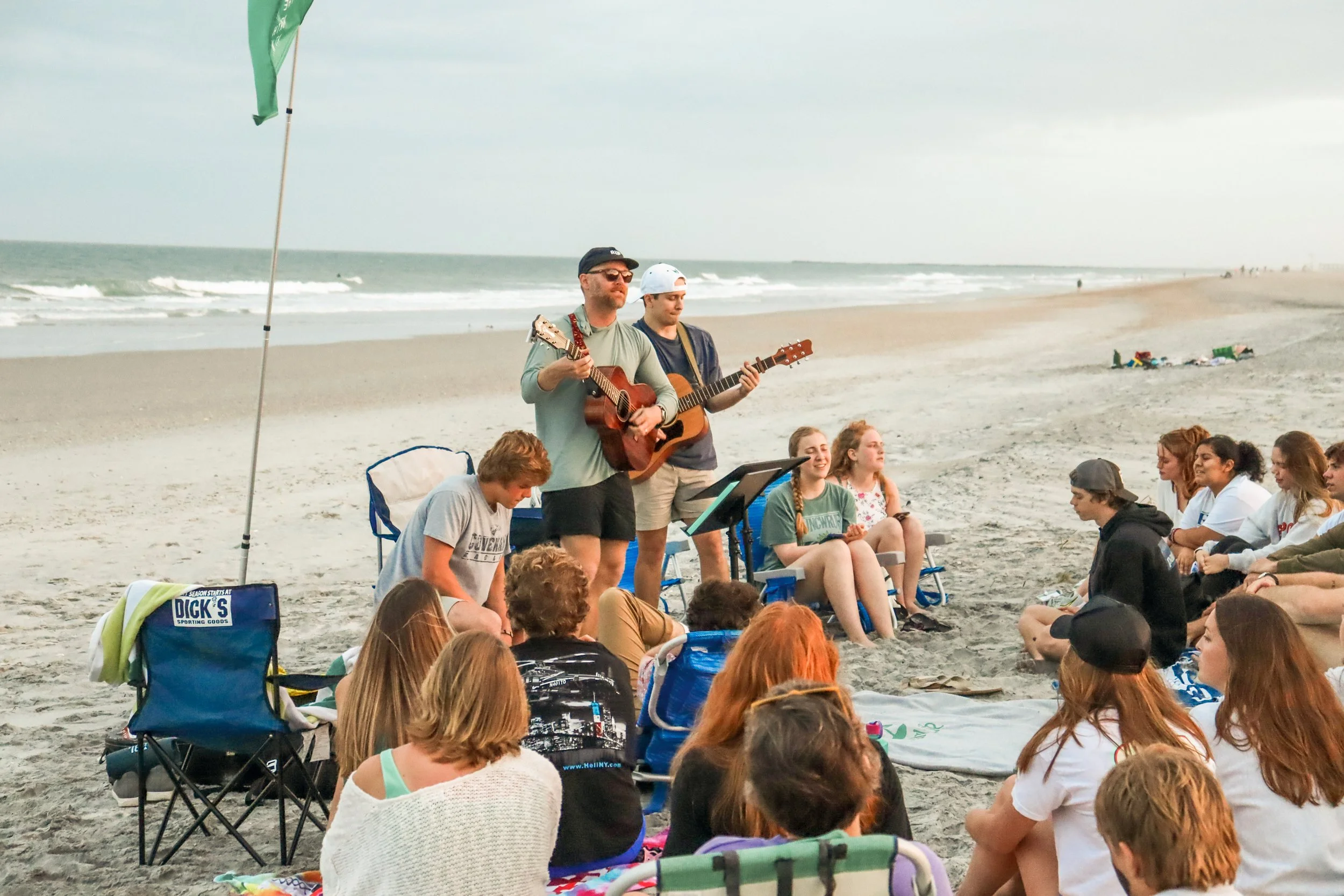 A group of people sitting on the beach watching two men perform with guitars.