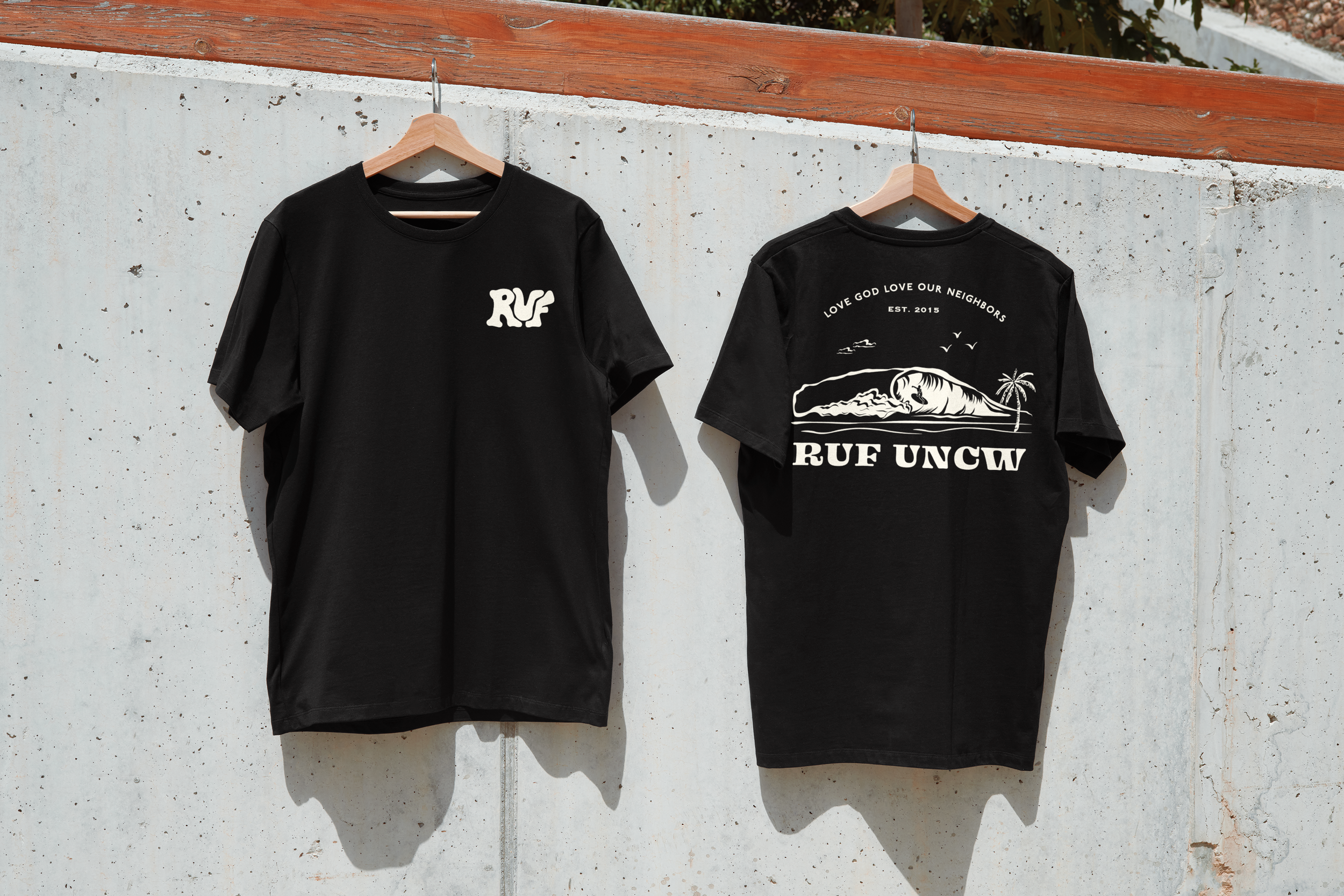 Two black T-shirts hanging on wooden hangers against a concrete wall, one front view and one back view. The front shirt has a small white logo that reads 'RUF' on the left chest. The back shirt features a white graphic of a raindrop on a landscape with a palm tree, birds, and words above and below the graphic that say 'LOVE GOD LOVE OUR NEIGHBORS EST. 2015' and 'RUF UNCW'.