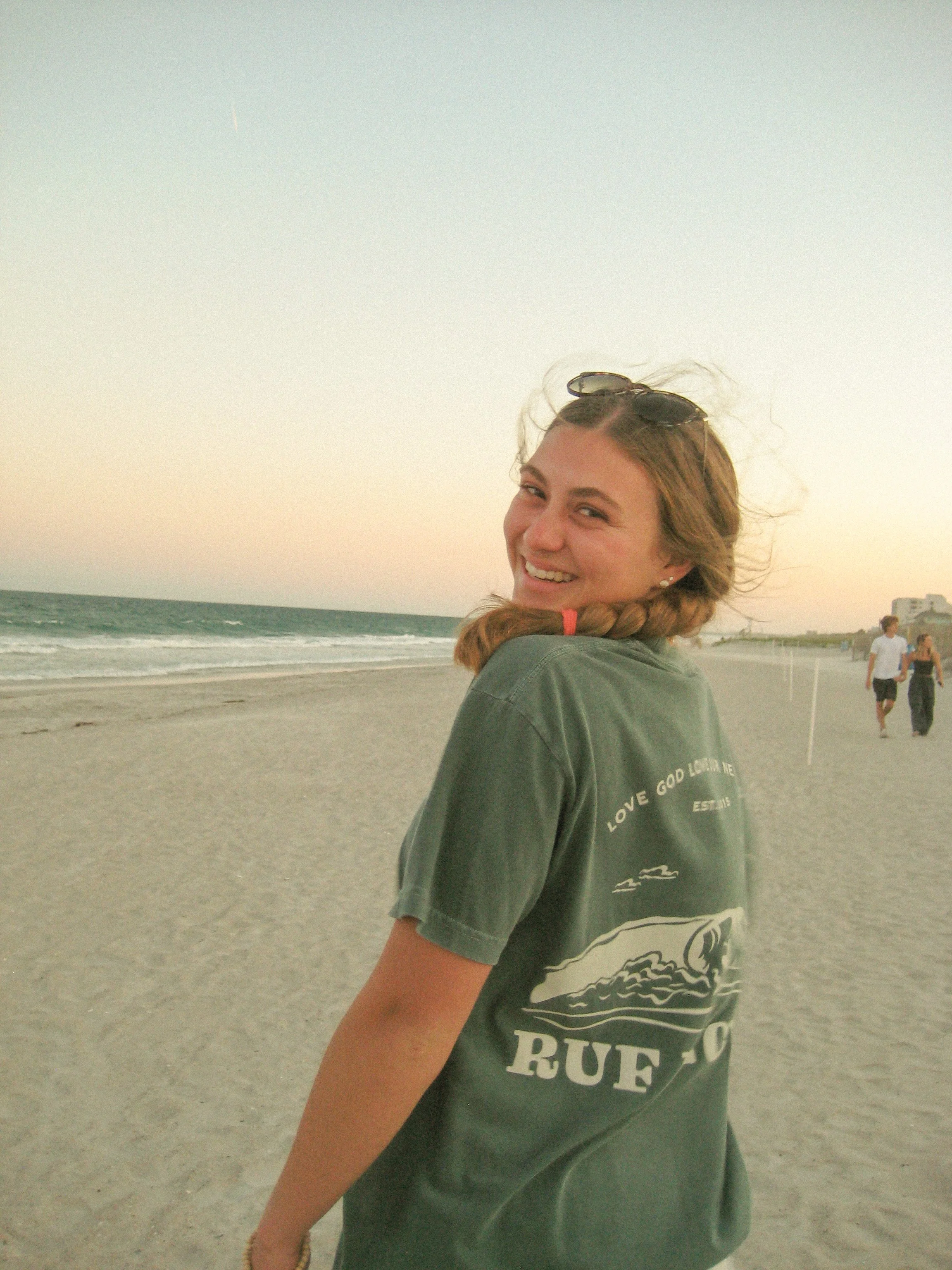 A young woman with sunglasses on her head, smiling at the camera, standing on a sandy beach at sunset.