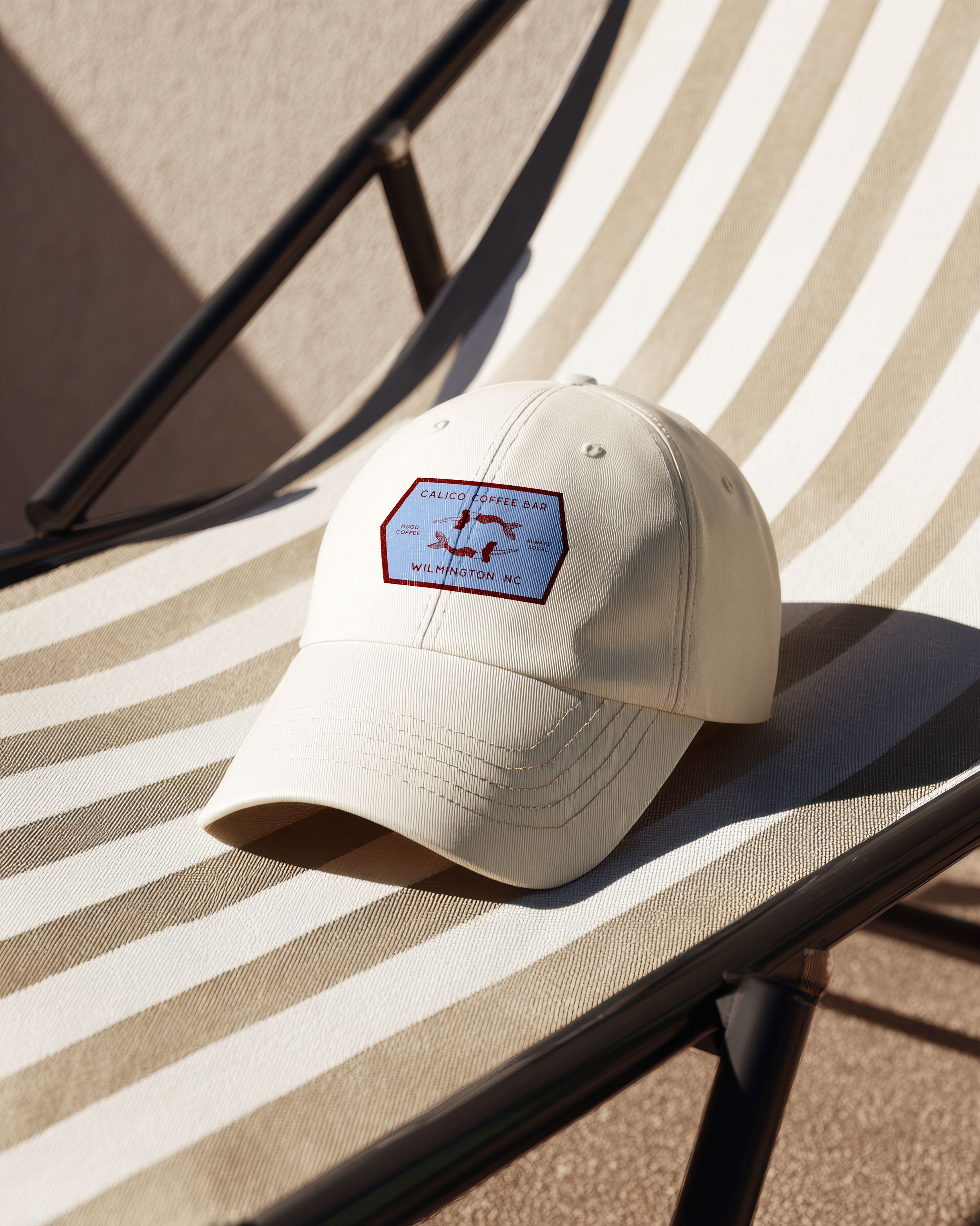 A white baseball cap with a logo sitting on a striped fabric surface, with a folding chair nearby.