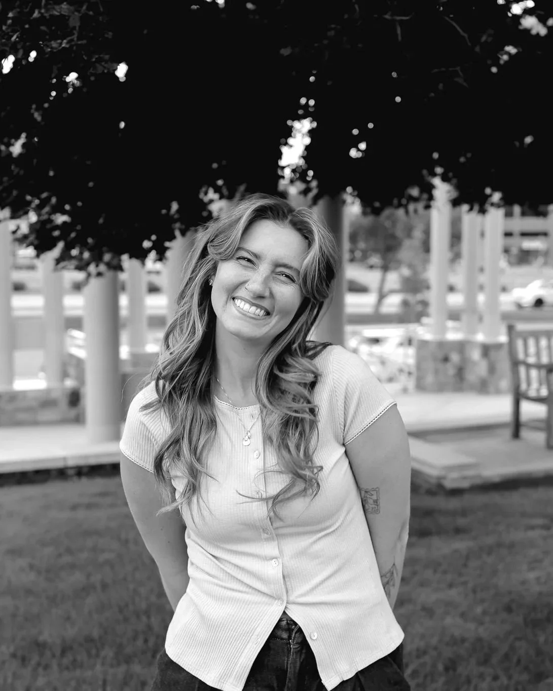 A woman smiling and posing outdoors with trees and a fence in the background, black and white photograph.