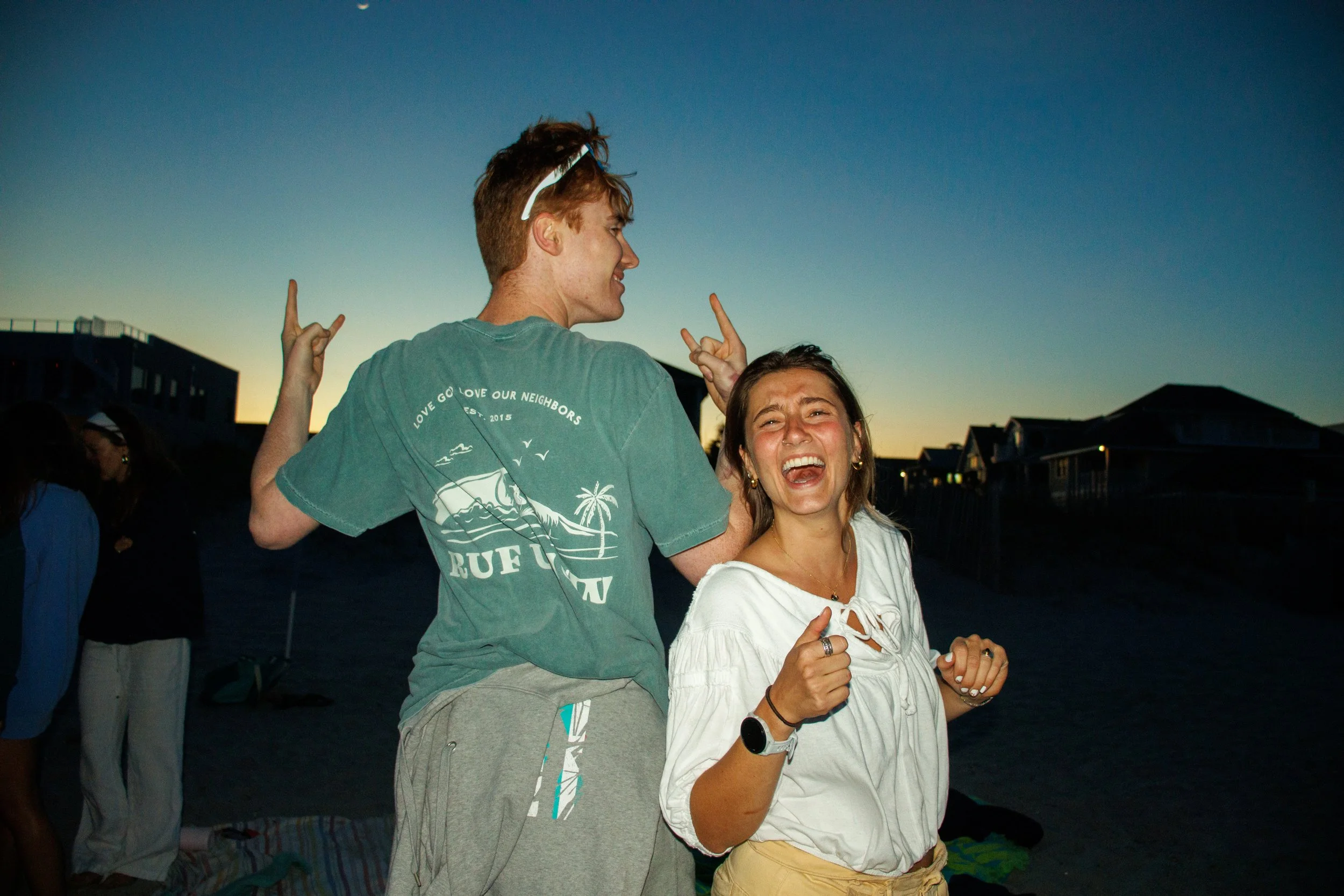 Two young people smiling and making peace signs at sunset on a beach, with houses in the background.