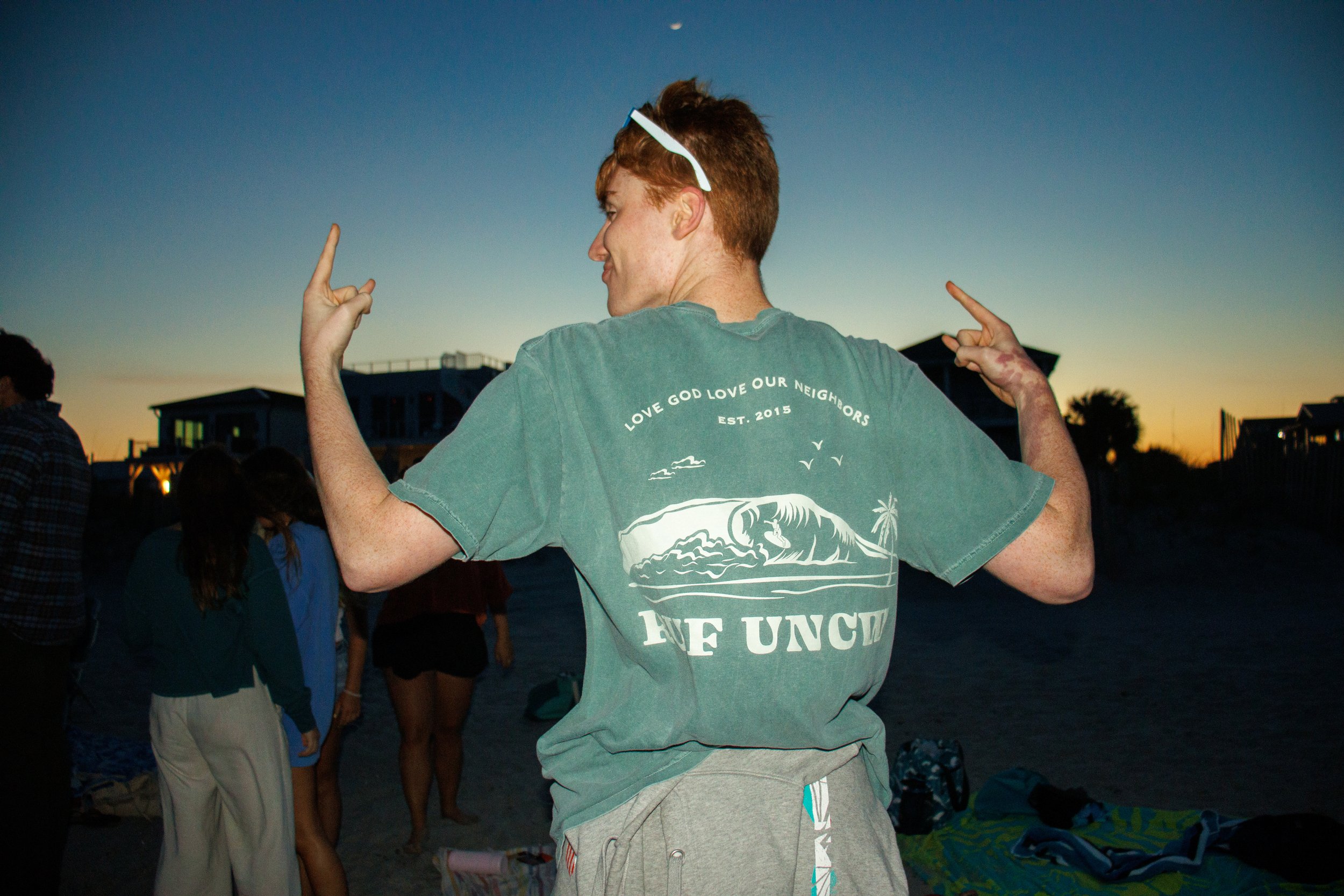 A young man with red hair and light skin standing on a beach at sunset, wearing a green t-shirt with a marine graphic and the text 'Love God Love Our Neighbors Est. 2015', and sunglasses on his head, making peace signs with both hands.