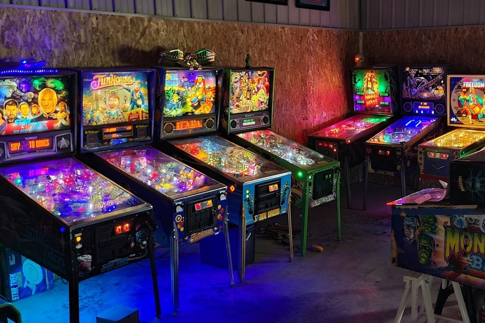 A row of vintage pinball machines with colorful lights, set against a wooden wall in a game room or arcade.