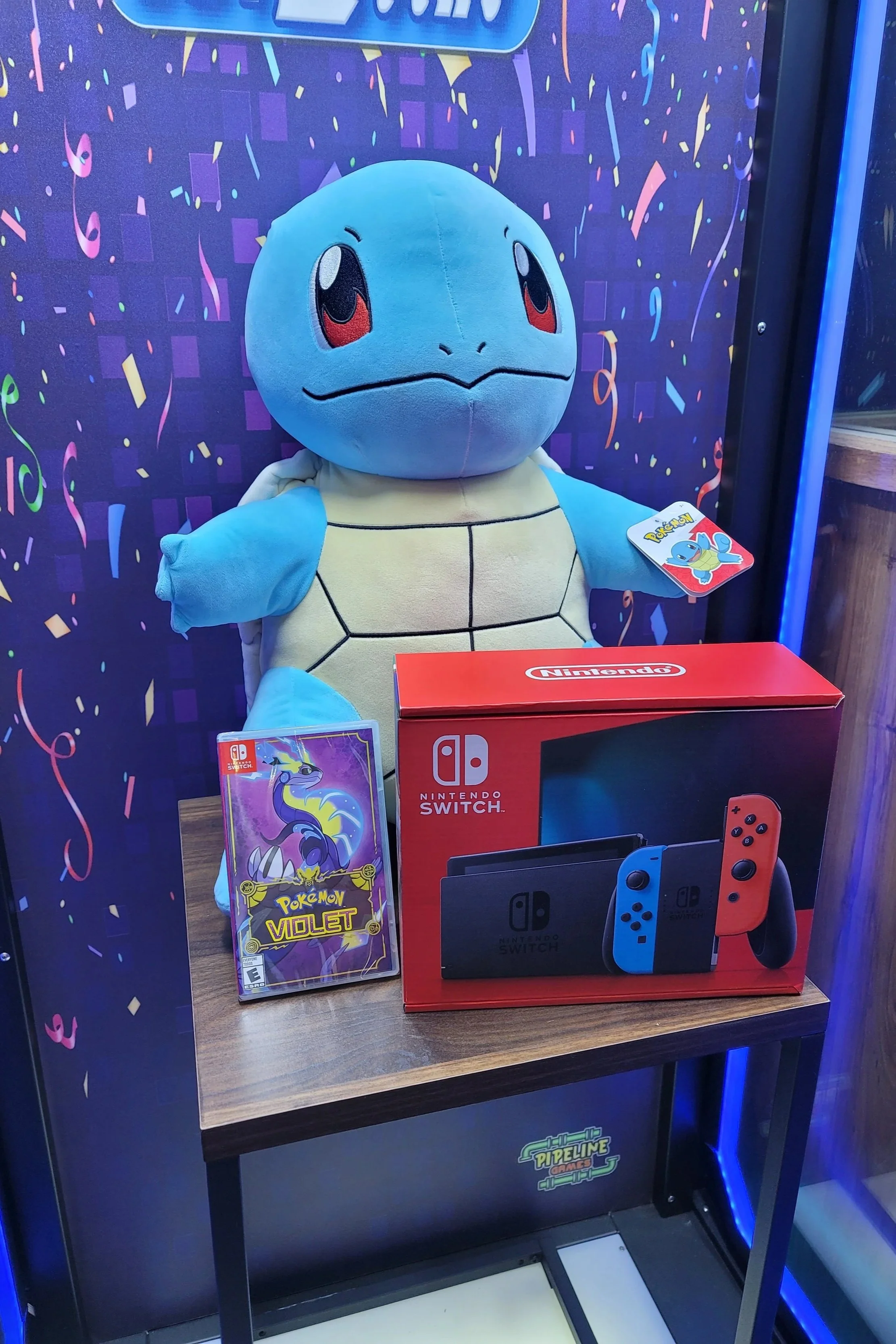 A plush Toy of Squirtle from Pokémon, a Pokémon Violet game case, and a Nintendo Switch box on a wooden table inside a gaming store with a colorful, confetti-patterned backdrop.