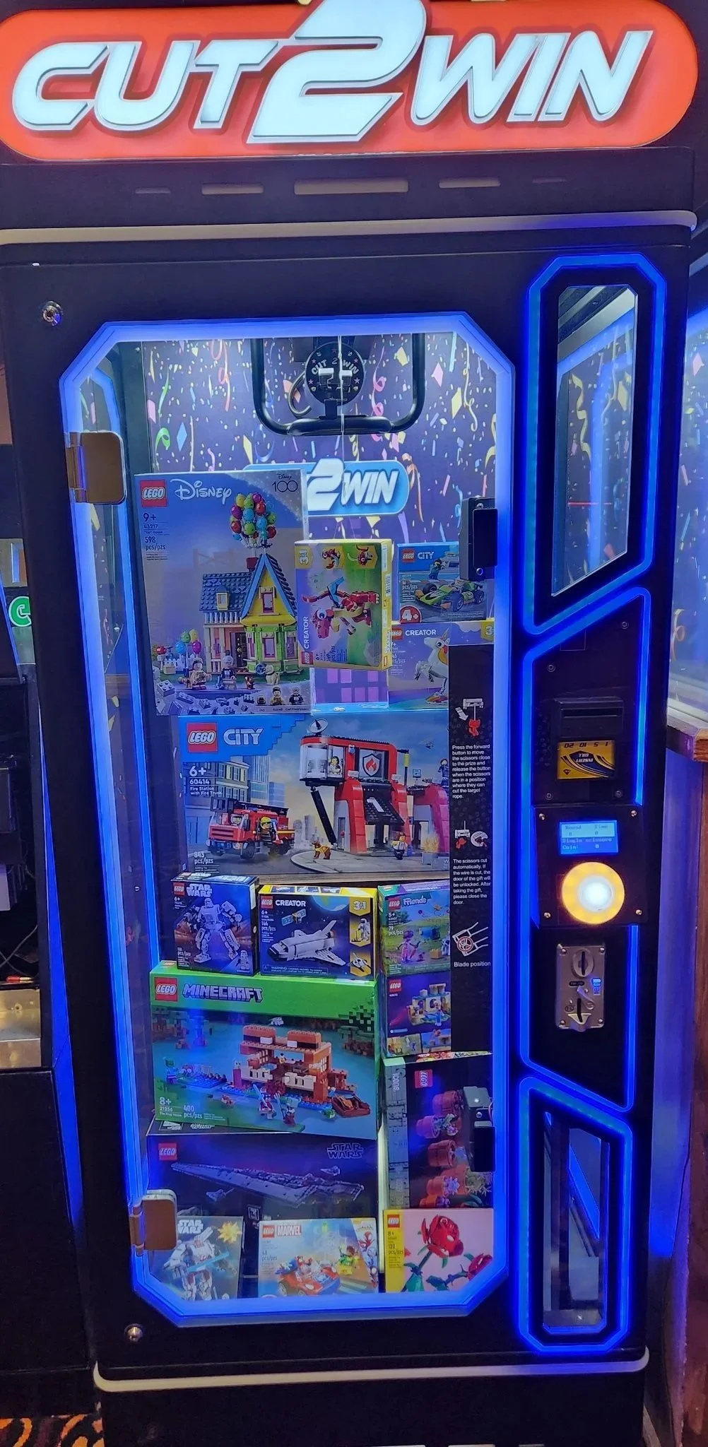 A claw machine filled with LEGO sets, including LEGO Disney, LEGO City, LEGO Minecraft, LEGO Star Wars, and Marvel sets, with a sign above reading 'CUTE 2 WIN'. The machine has a blue LED rim and a control panel with a joystick and buttons.