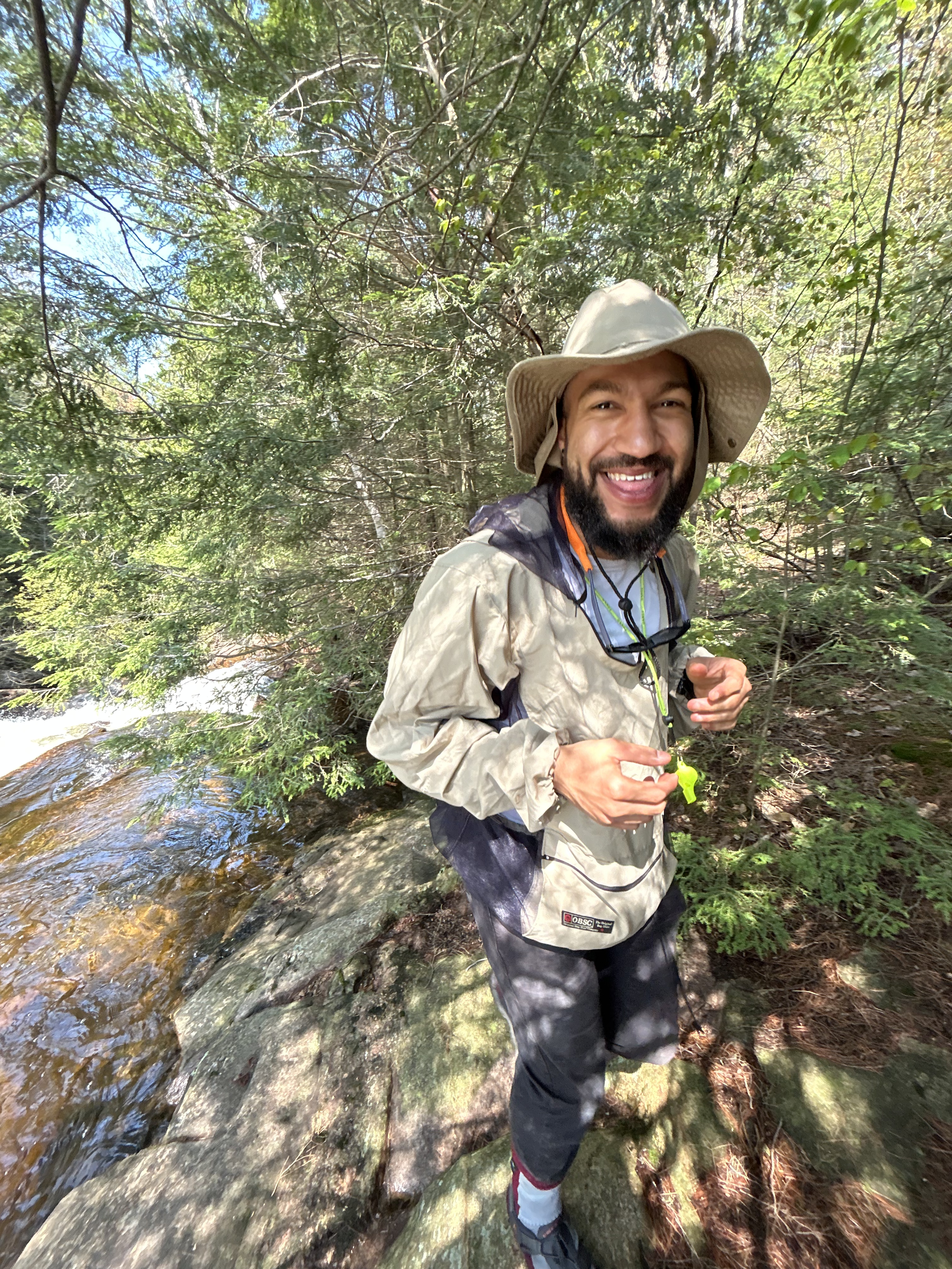 A man smiling and dressed in outdoor hiking gear standing on rocks beside a small stream in a wooded area.