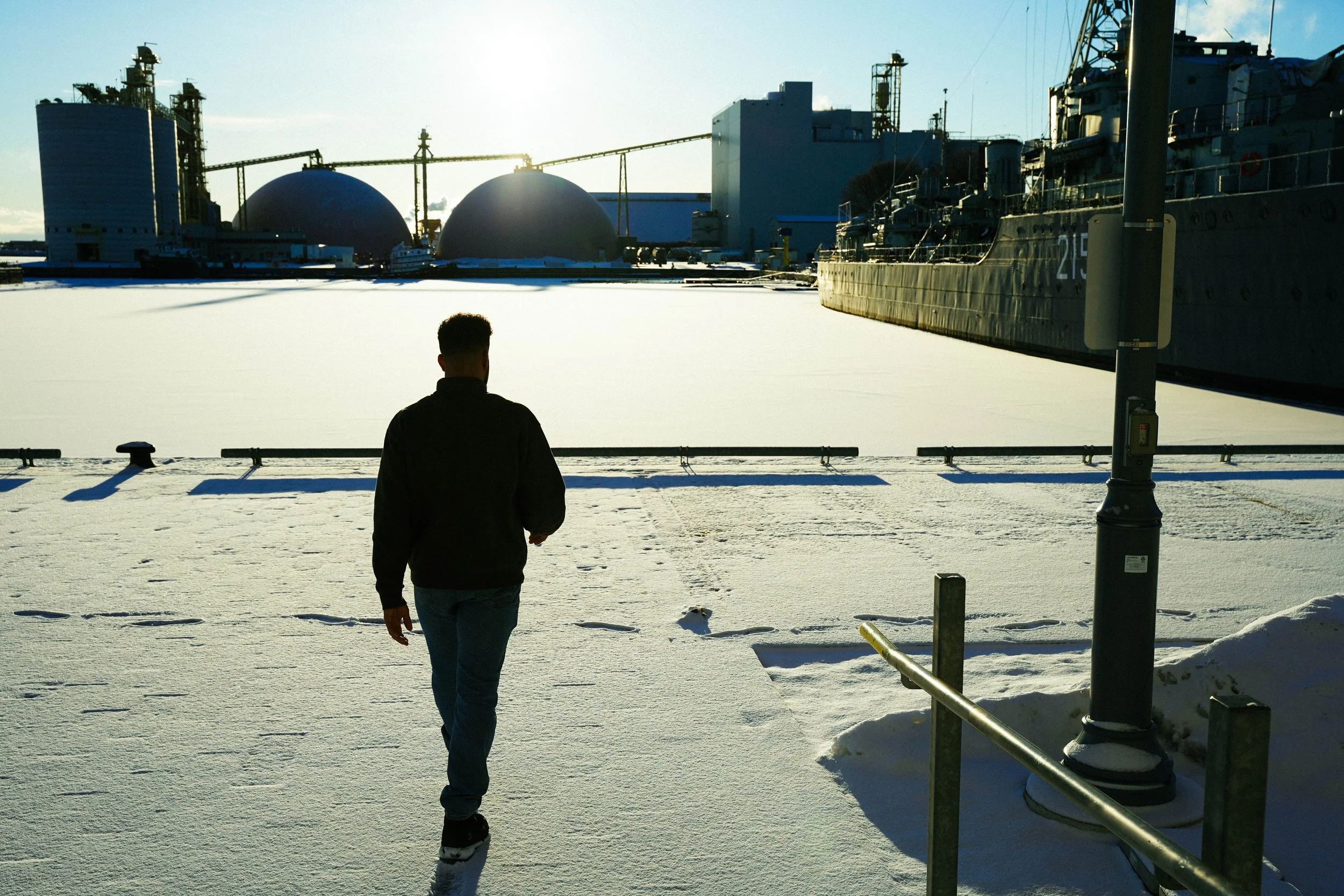 A person walking in a snowy port area with ships and industrial structures in the background, during sunset.