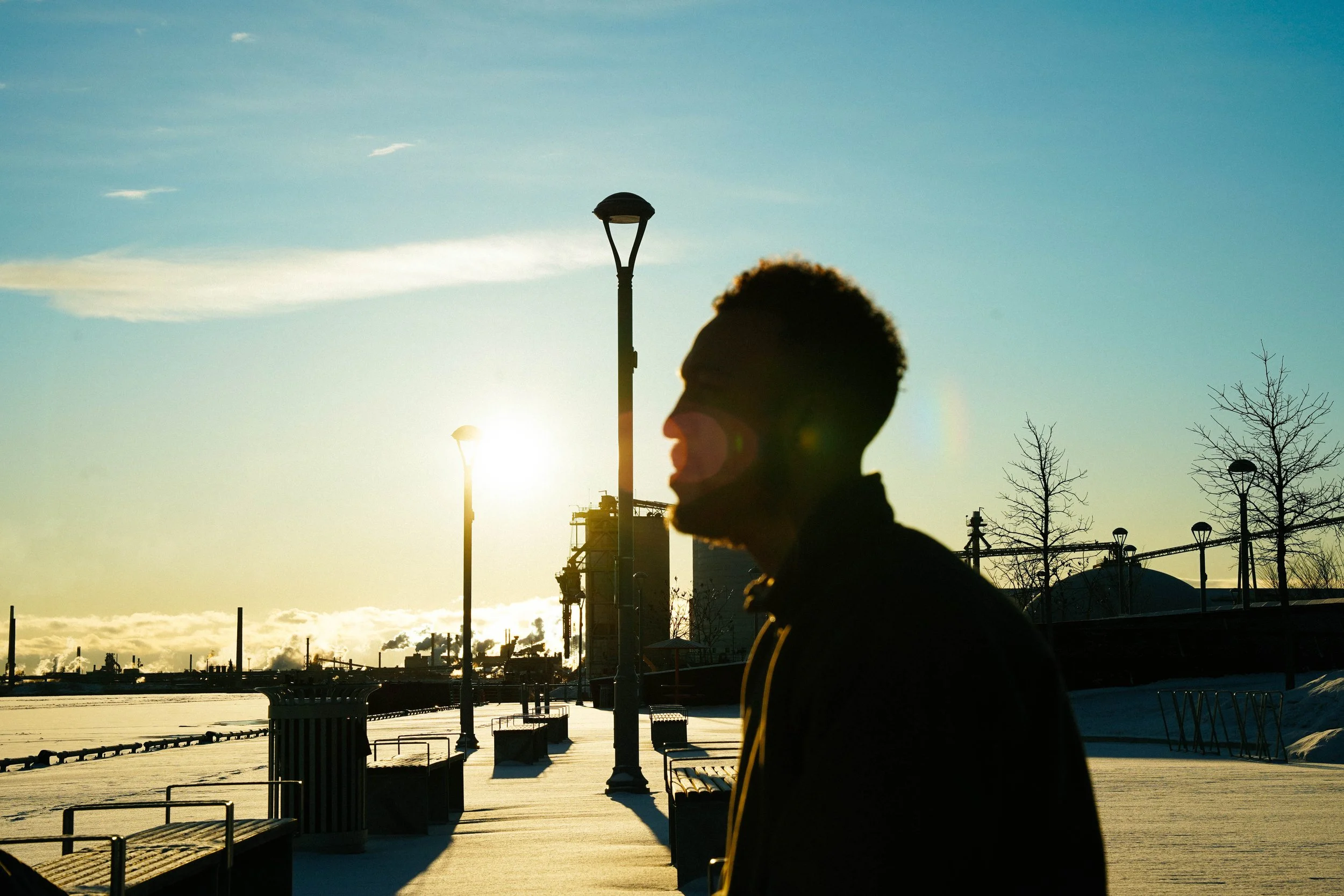 A silhouette of a man with short curly hair and glasses, walking along a snow-covered promenade during sunset, with industrial chimneys and leafless trees in the background.