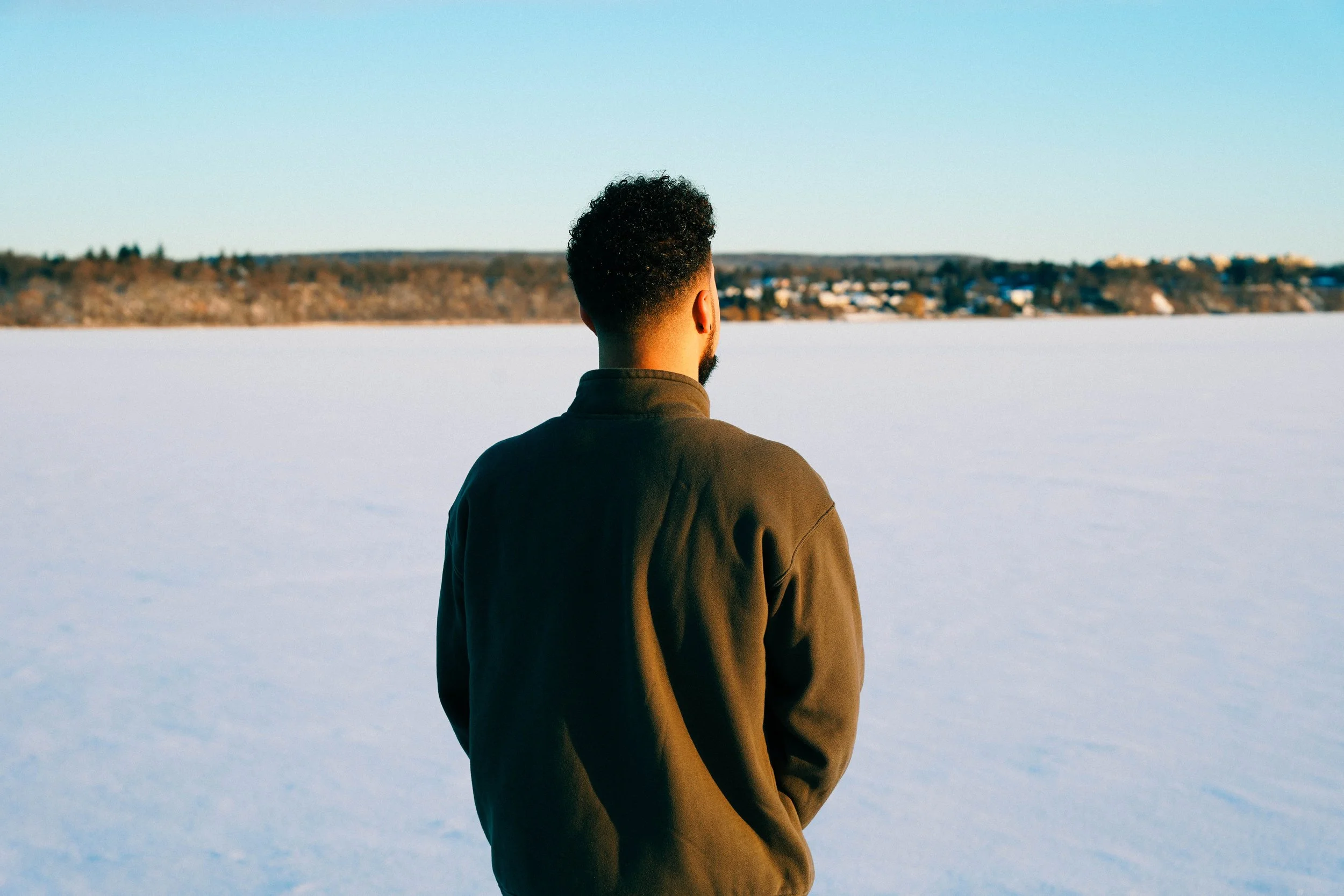 A man with curly hair and a beard stands with his back to the camera, looking across a frozen lake or body of water, with a distant shoreline and houses visible under a clear blue sky.