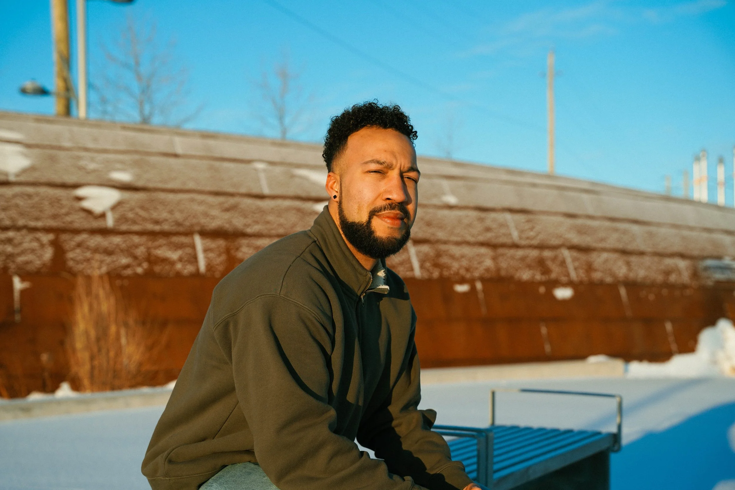 A man sitting on a bench outdoors in a snowy area with a fence and a brick wall behind him, under a clear blue sky.