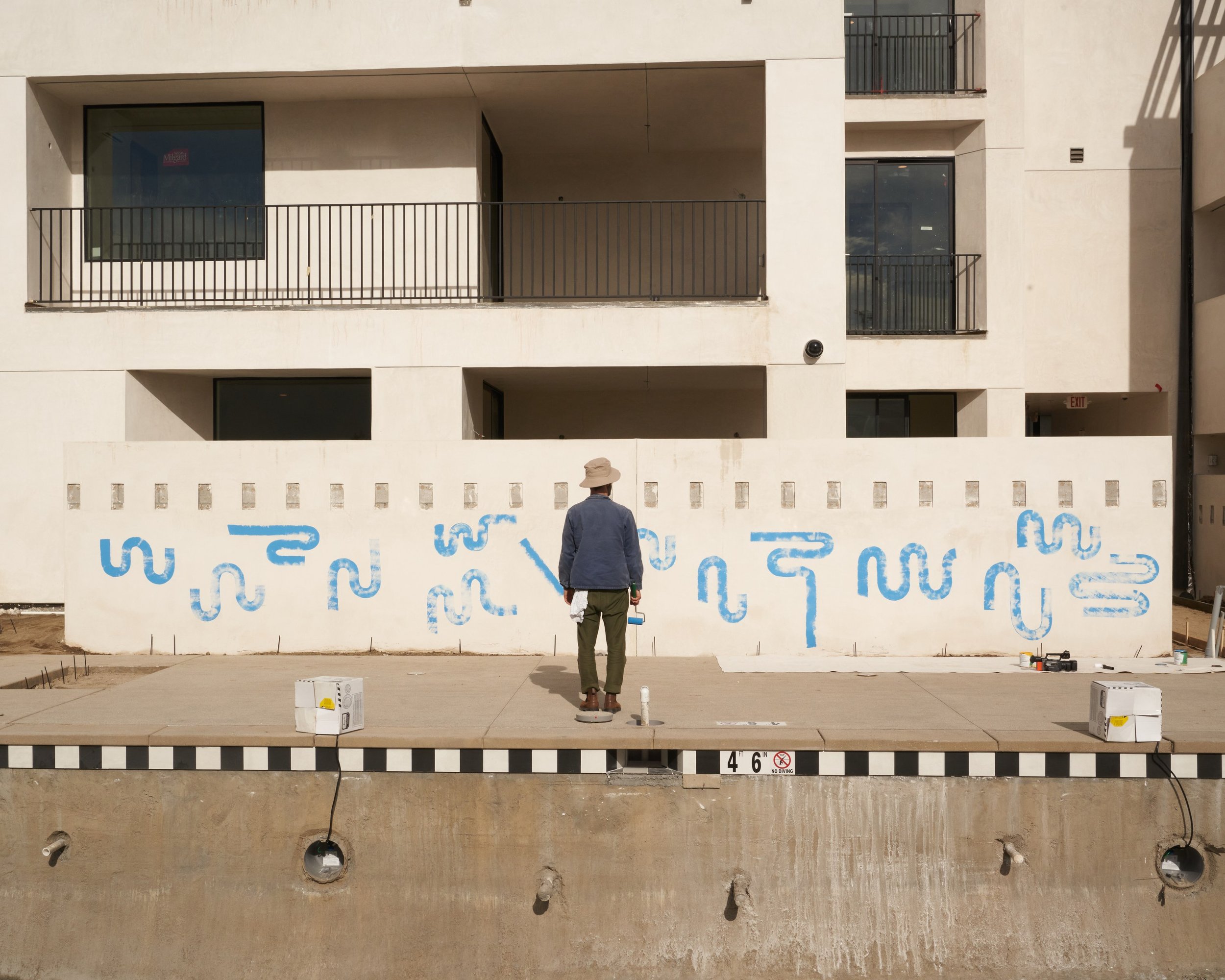 Sean W Spellman painting a blue abstract mural on a courtyard wall beside a pool under construction at Drift hotel in Palm Springs, CA