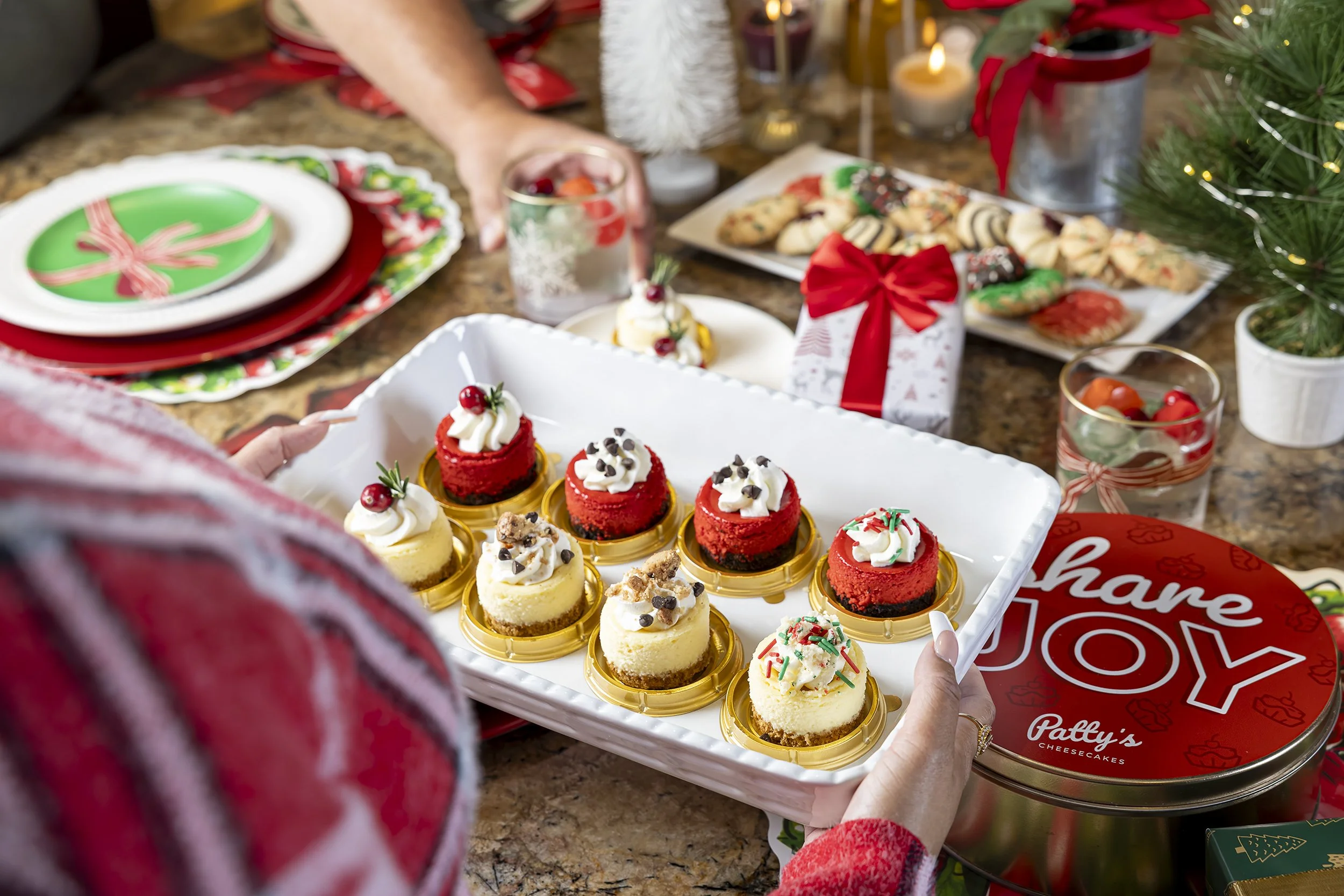 Person holding a tray of assorted mini cheesecakes topped with whipped cream and holiday decorations at a festive Christmas gathering.