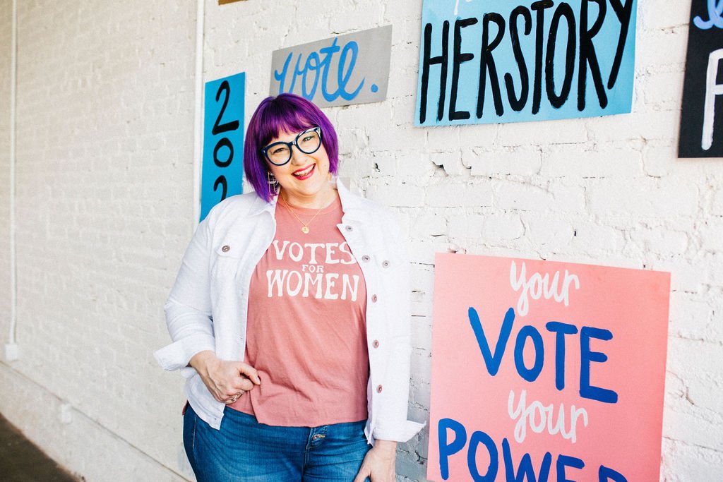 Woman with purple hair, glasses, pink shirt reading 'Votes for Women', white jacket, and blue jeans standing next to campaign signs promoting voting and women's rights against a white brick wall.