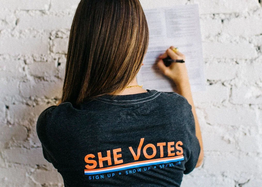 Back of a woman with brown hair wearing a black t-shirt that says "SHE VOTES" and additional text promoting voter registration, standing in front of a white brick wall.