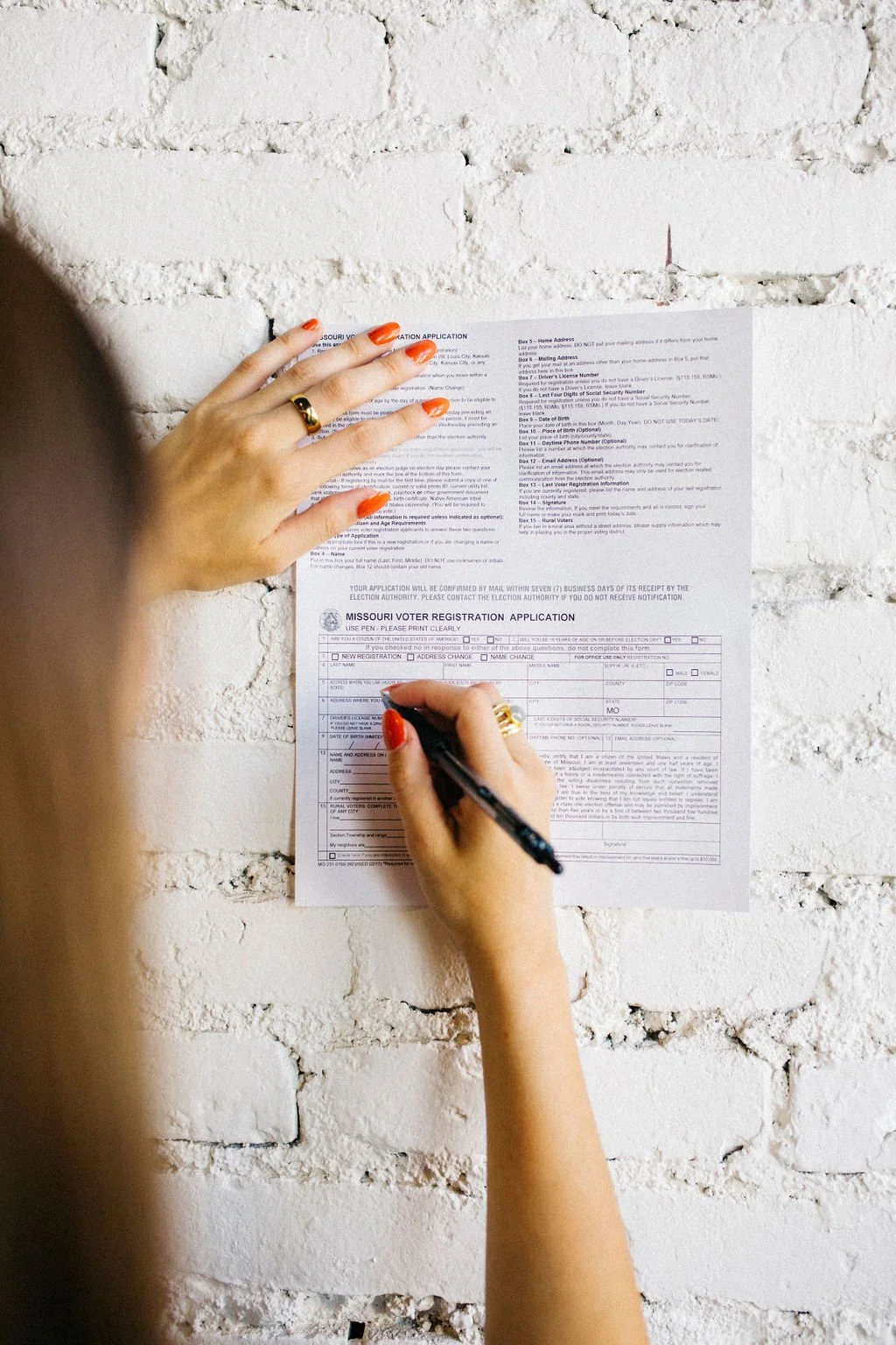 Person filling out a Missouri voter registration application form on a white brick wall.