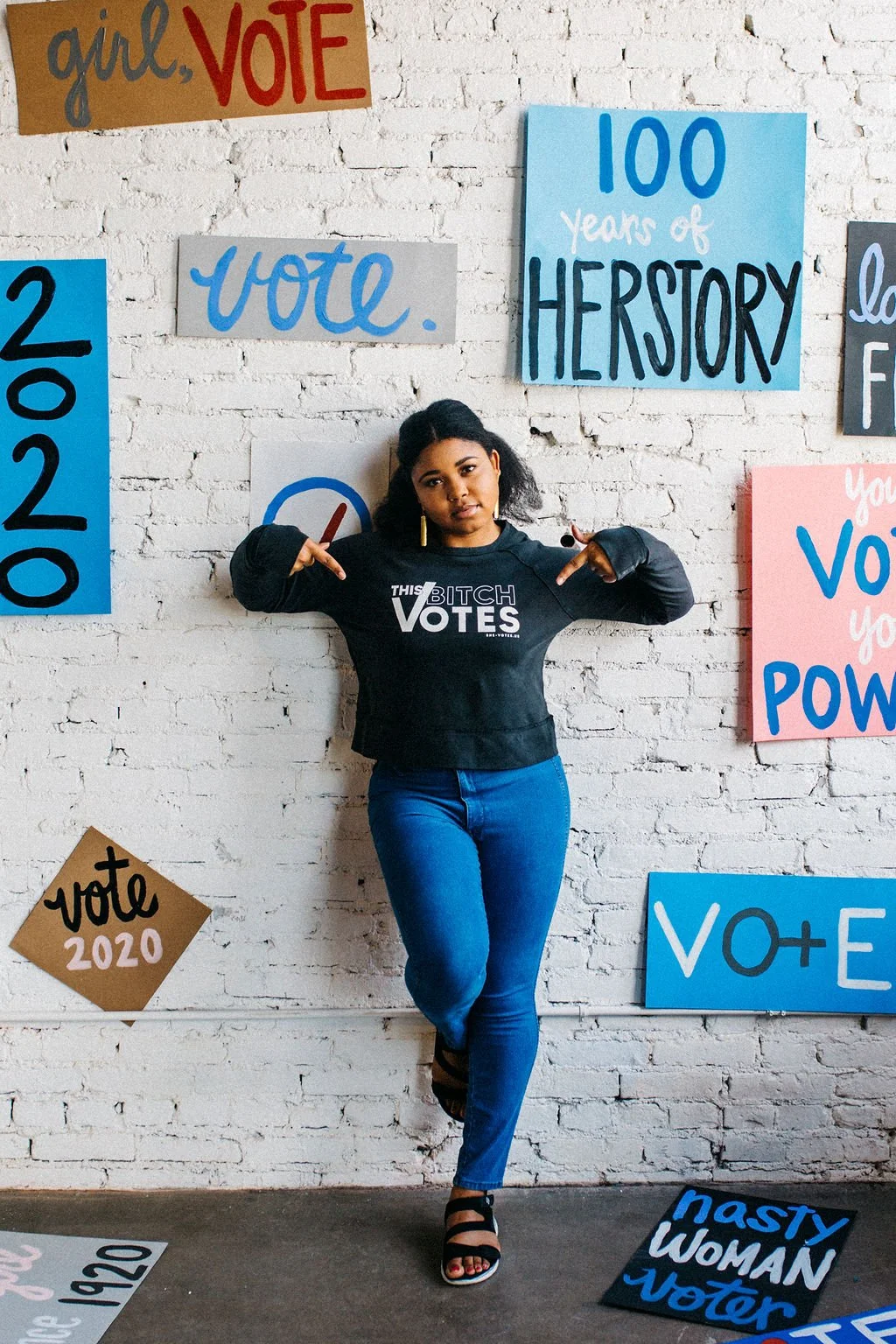 A woman standing against a white brick wall decorated with colorful signs promoting voting and history, wearing a black sweatshirt with the words "This Bitch VOTES," and pointing at the sweatshirt.