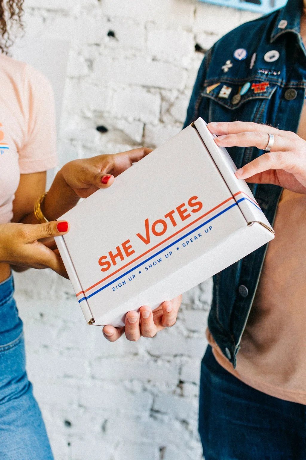 Two women exchanging a white box with orange and blue text that reads 'SHE VOTES' and a message about signing up, showing up, and speaking up. One woman has red nail polish and gold jewelry, the other wears a denim vest with pins. They stand in front