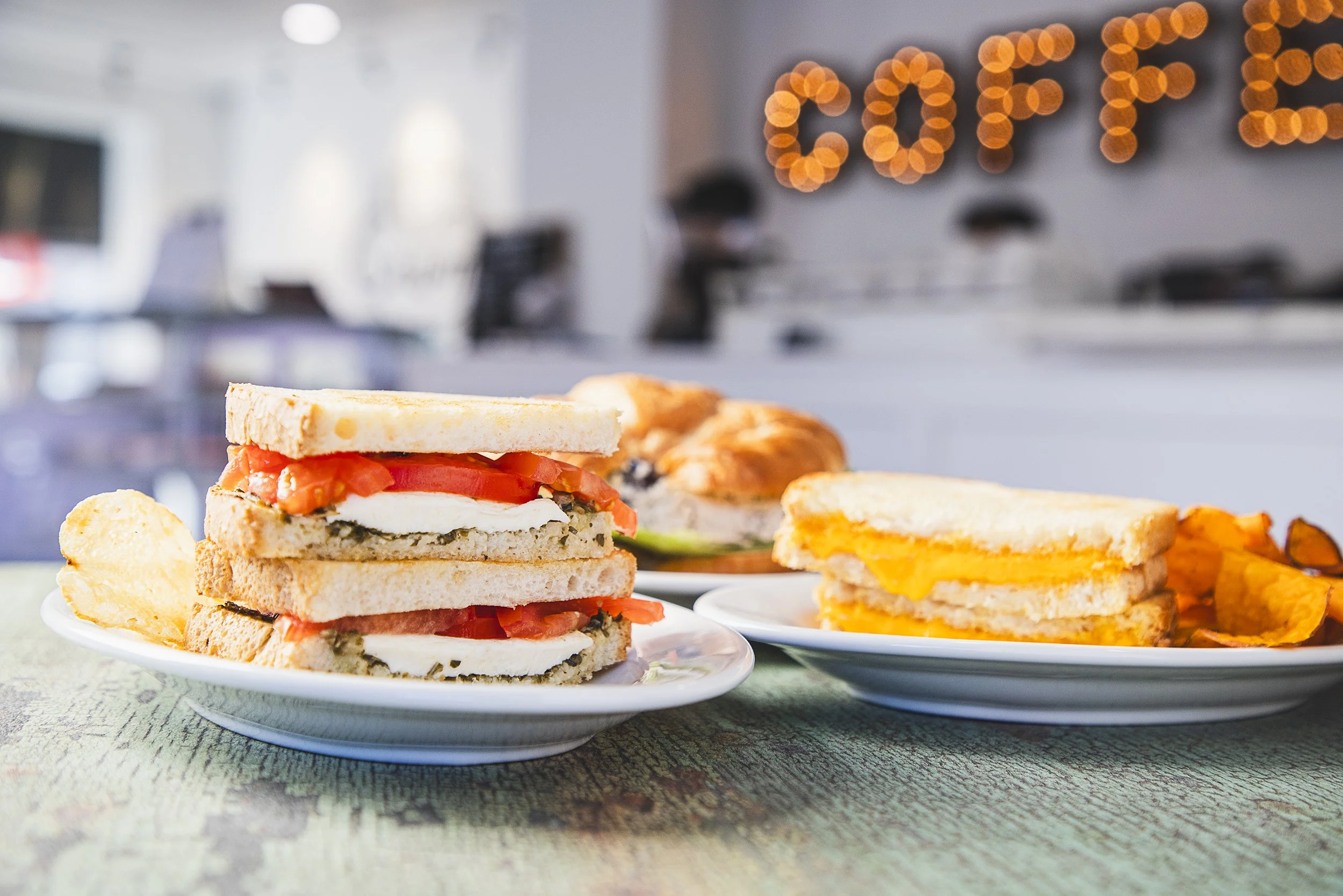 Close-up of a plate with a triple-layer sandwich containing tomatoes, cheese, and herbs, accompanied by potato chips. In the background, there is another sandwich with melted cheese, a croissant sandwich, and chips, all on white plates in a café with