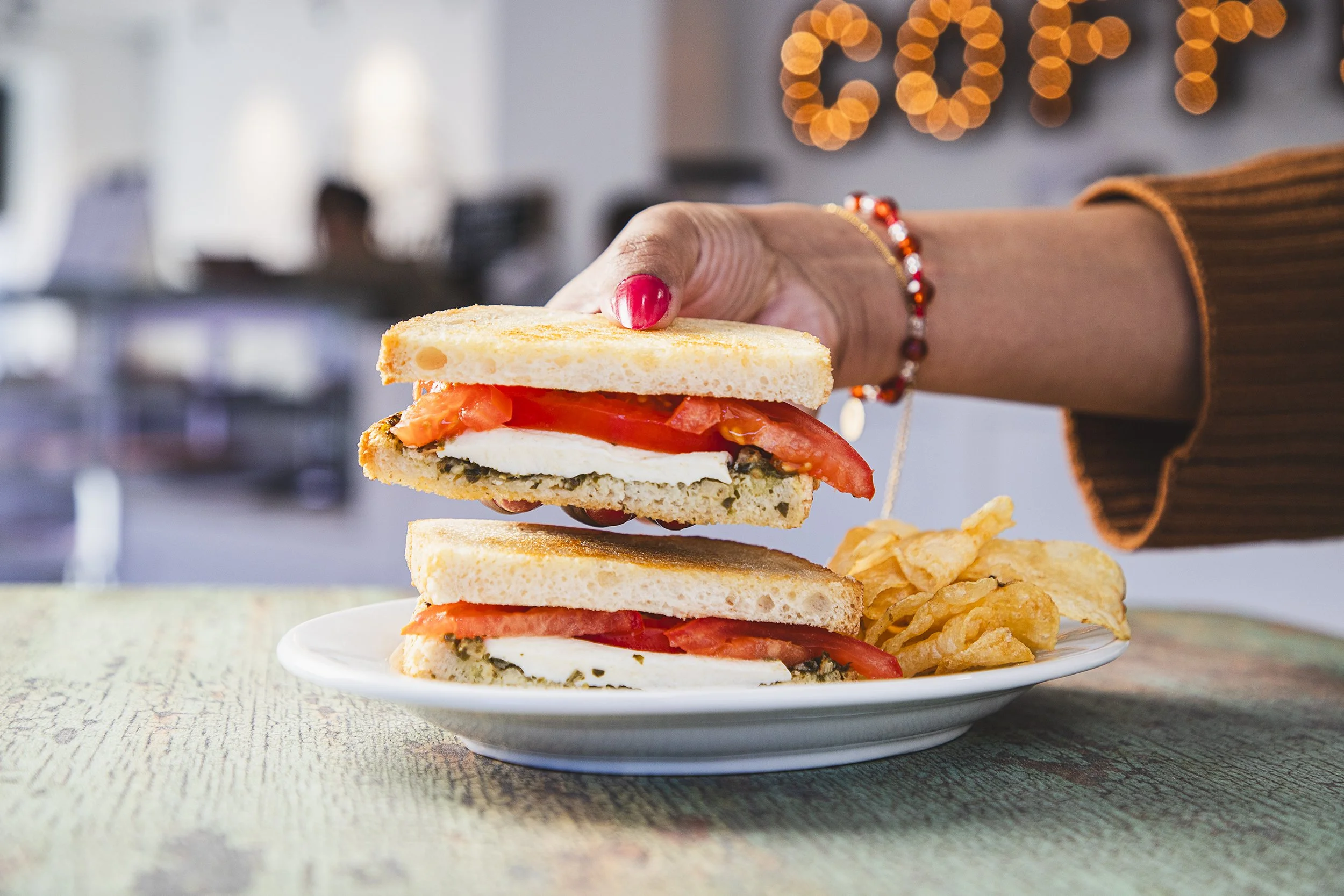 A person holding a sandwich filled with tomato, mozzarella cheese, and pesto, with potato chips on a plate on a wooden table.